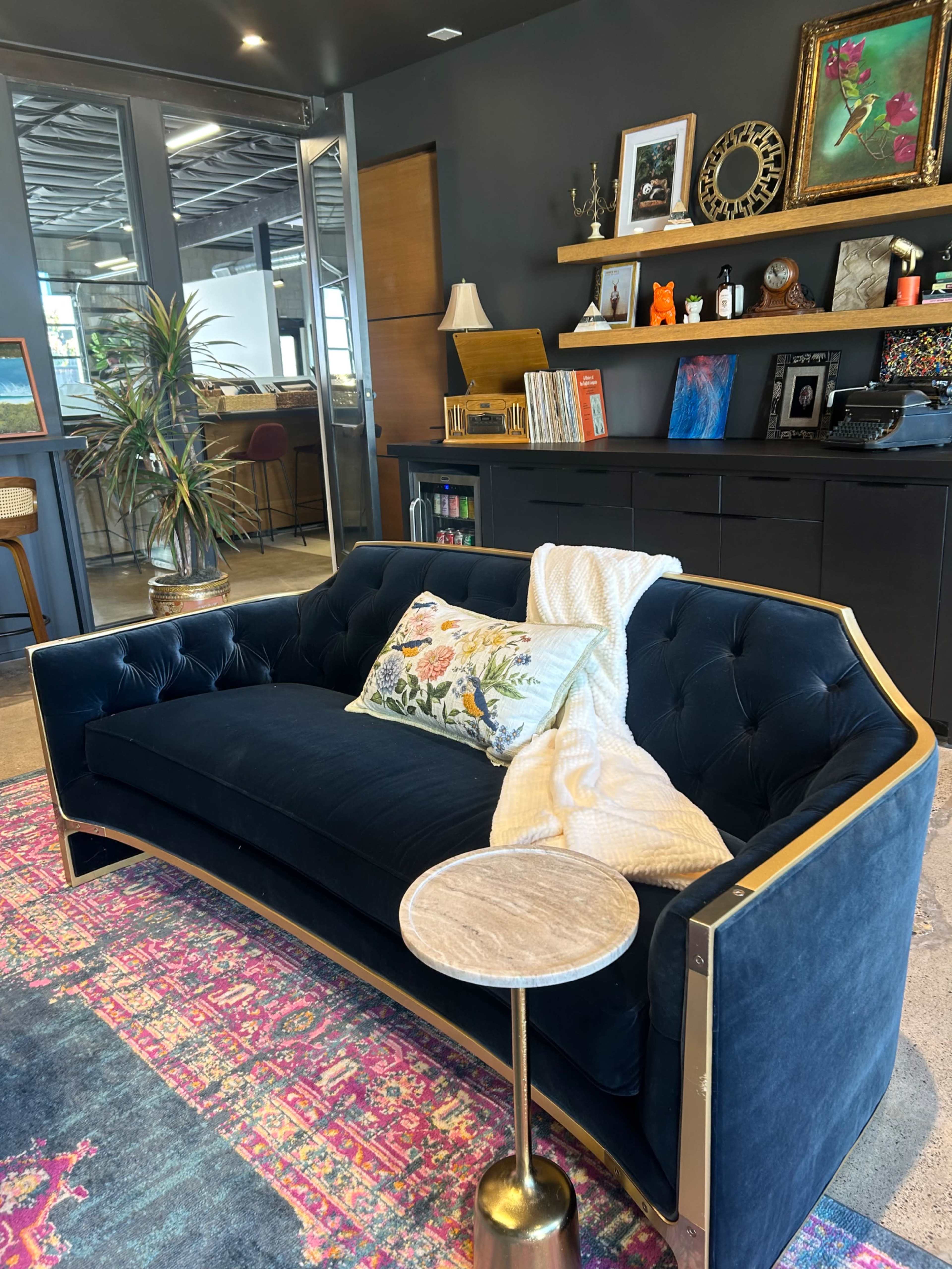 A dark blue tufted couch is positioned in a stylish interior, accompanied by a small golden side table and decorative elements on the wall and shelves.