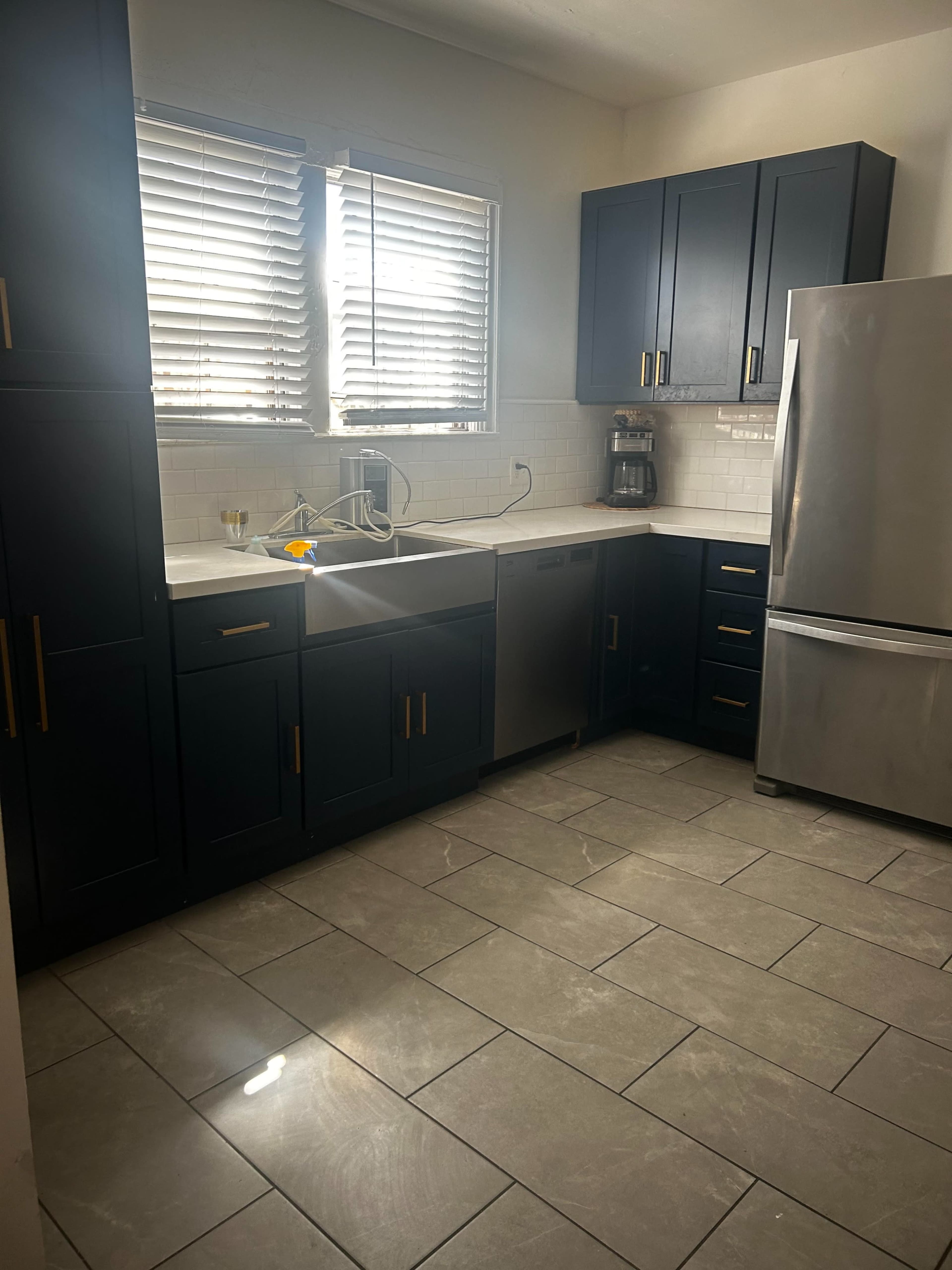 The image shows a modern kitchen featuring dark blue cabinets, a stainless steel refrigerator, and a white countertop with a sink and window above it.