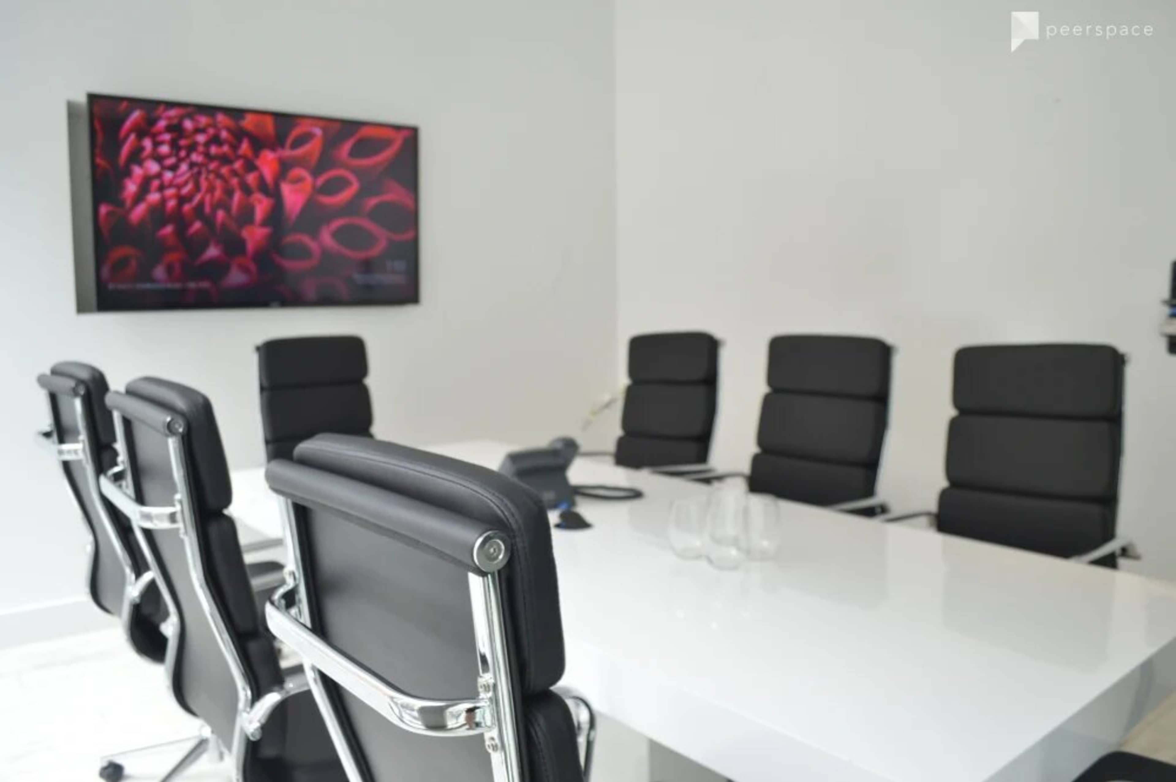 A conference room is set up with a large table, six black leather chairs, and a television mounted on the wall displaying a colorful image.