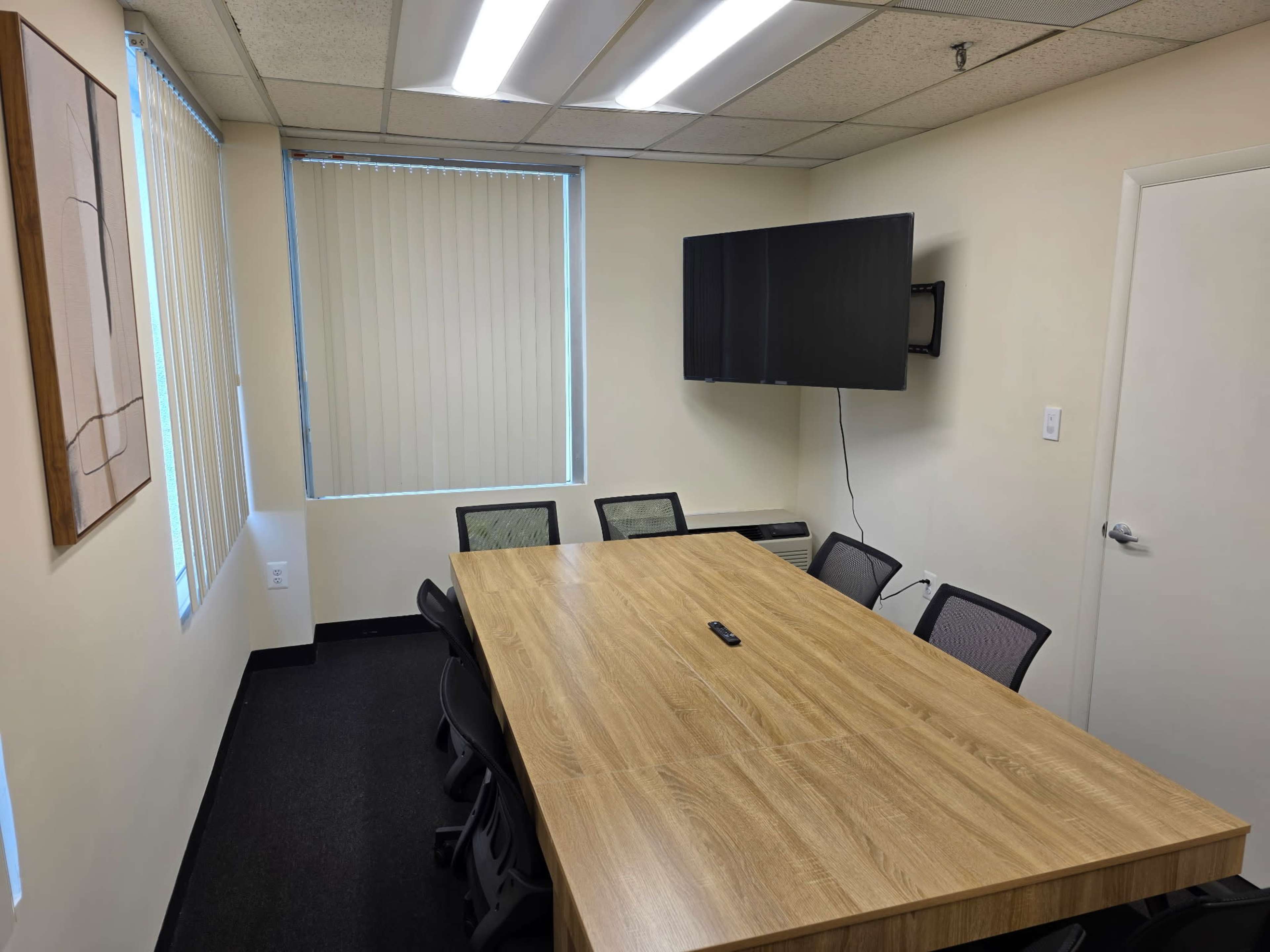 The image shows a conference room with a large wooden table, black chairs, a wall-mounted television, and windows covered with vertical blinds.