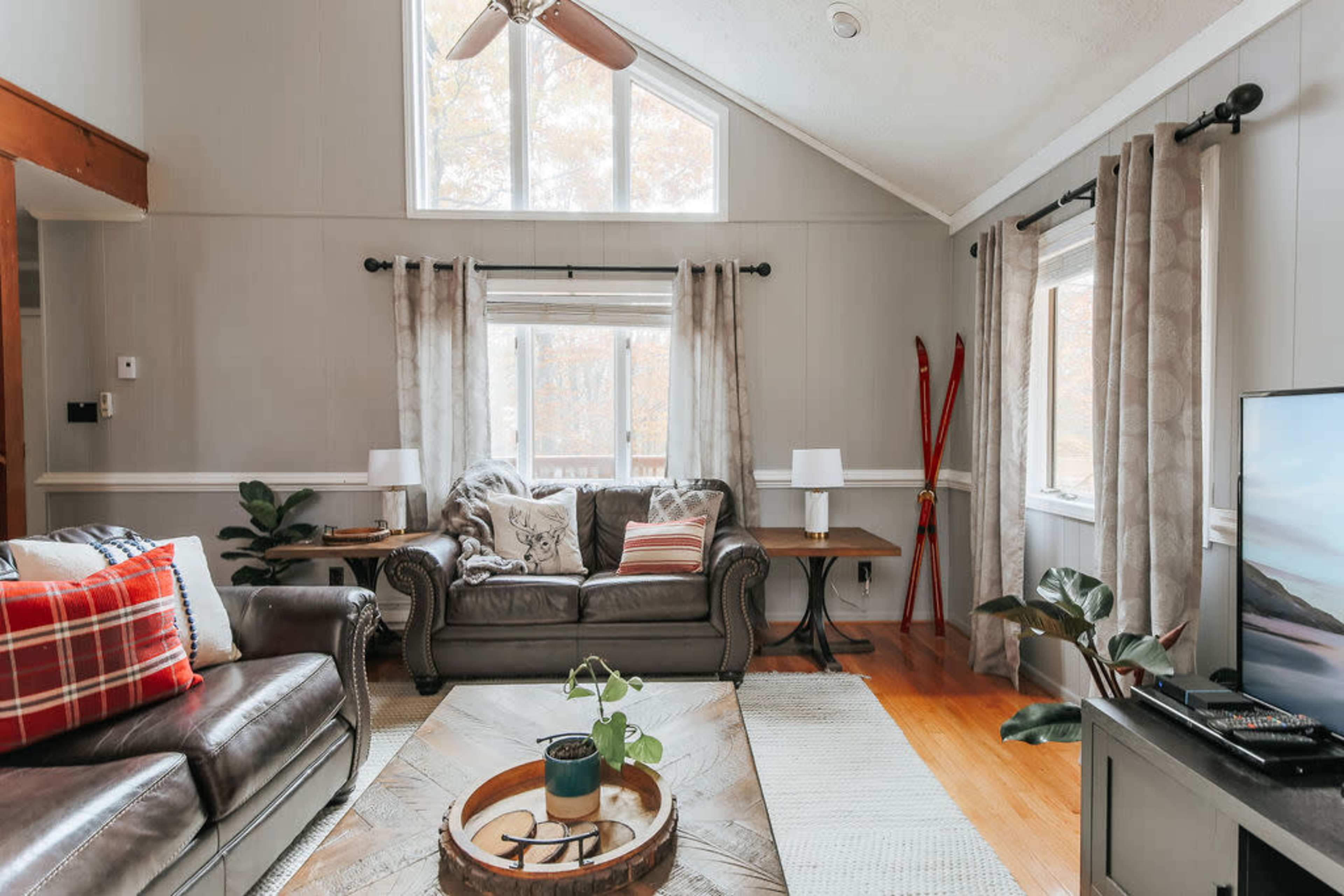 The living room features a high, sloped ceiling with large windows, a leather sofa, a wooden coffee table, and decorative elements like plants and ski equipment.