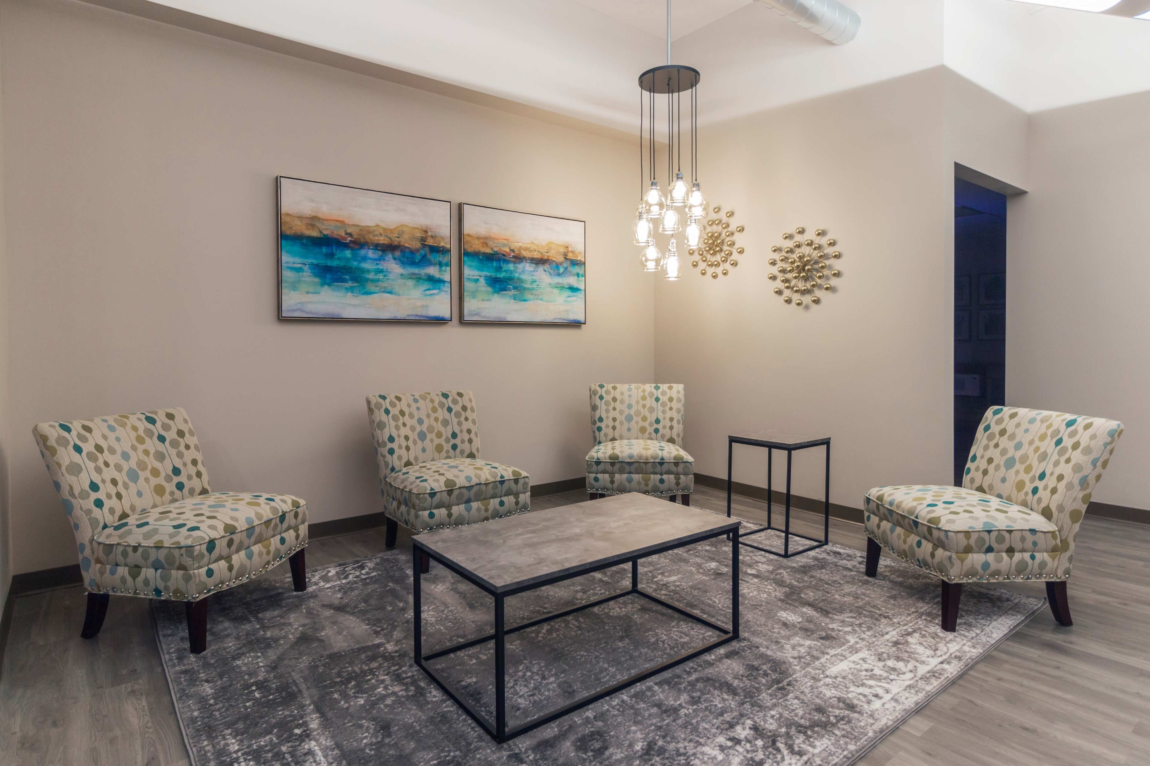 A modern waiting area with four patterned chairs arranged around a central coffee table on a rug, illuminated by a pendant light.