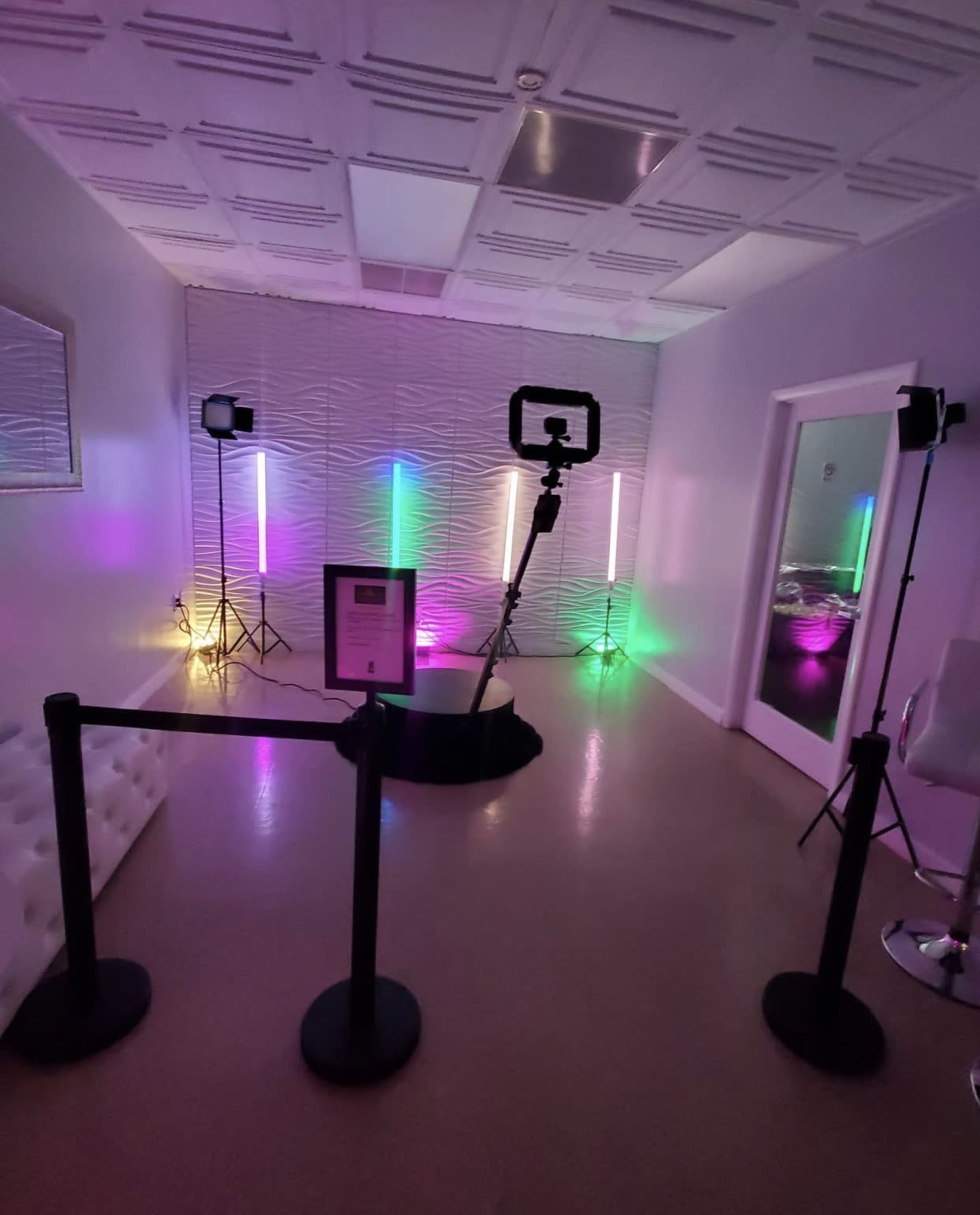 The image shows a modern room with colorful lights, a tripod with a ring light, and a velvet rope barrier separating the setup from the rest of the space.