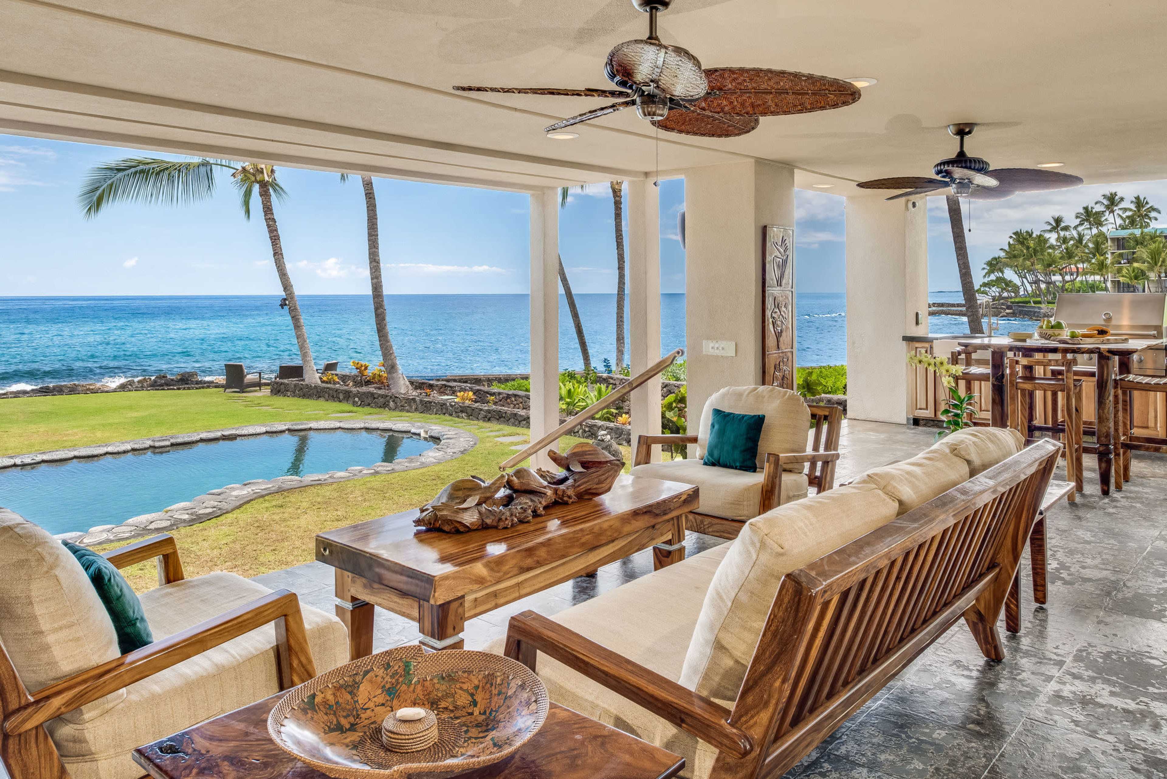 A well-furnished patio overlooking a pool and the ocean, with palm trees in the background.