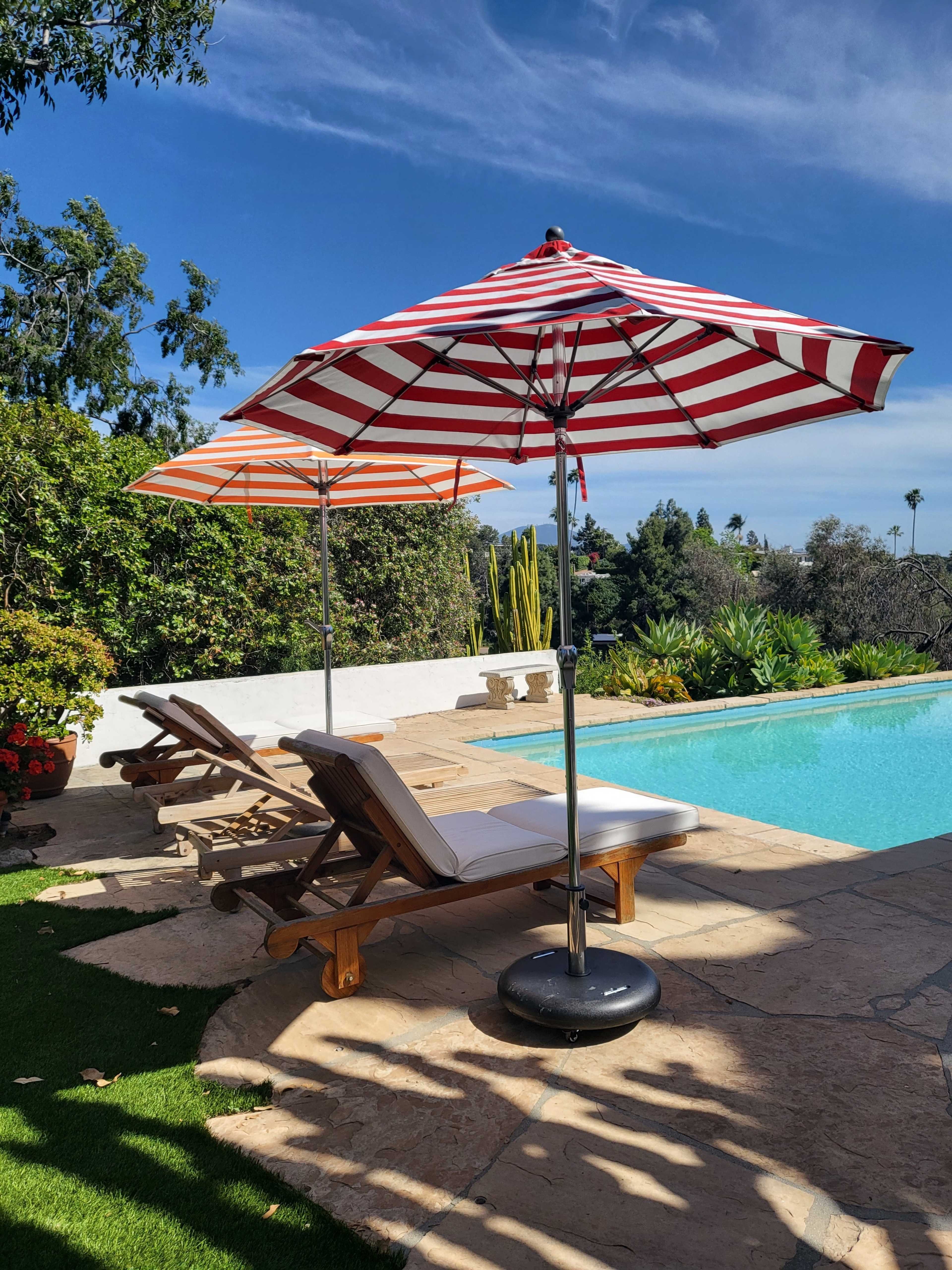 Two lounge chairs with striped umbrellas are positioned beside a swimming pool surrounded by greenery and stone patio.