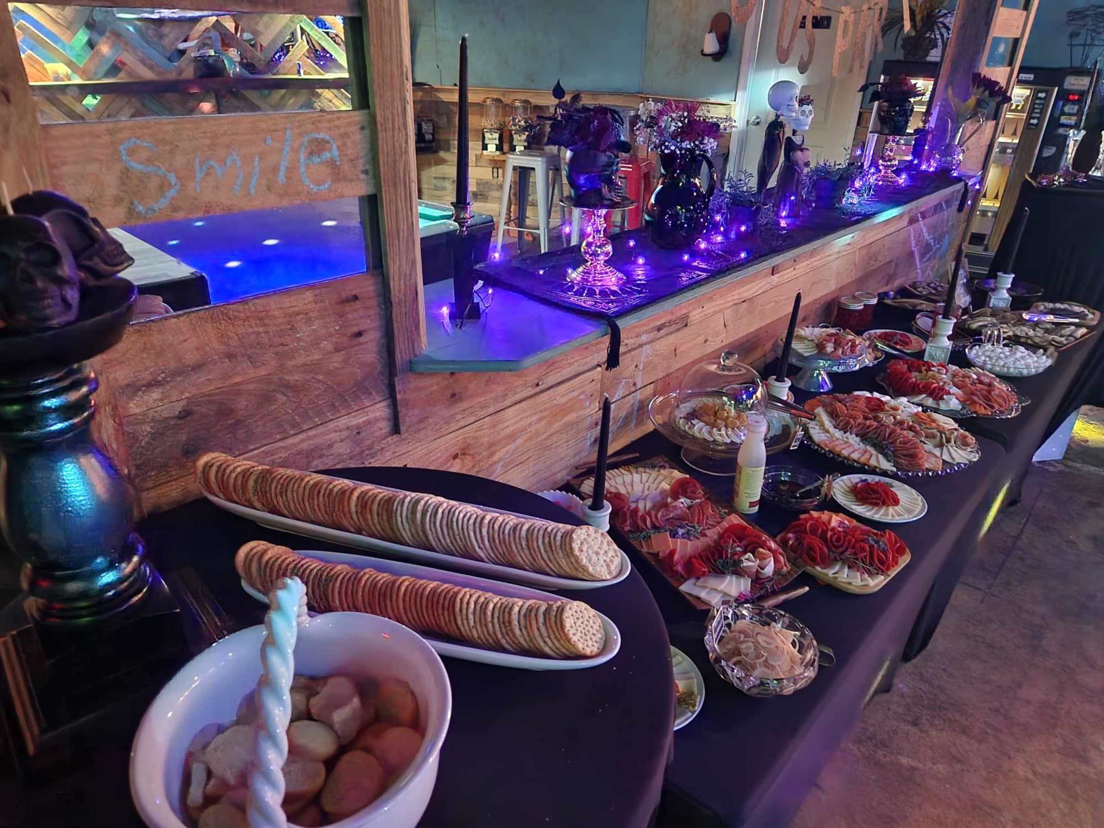 A variety of food items, including cookies, seafood, and pasta, are displayed on a table decorated with purple lighting and Halloween-themed decor.