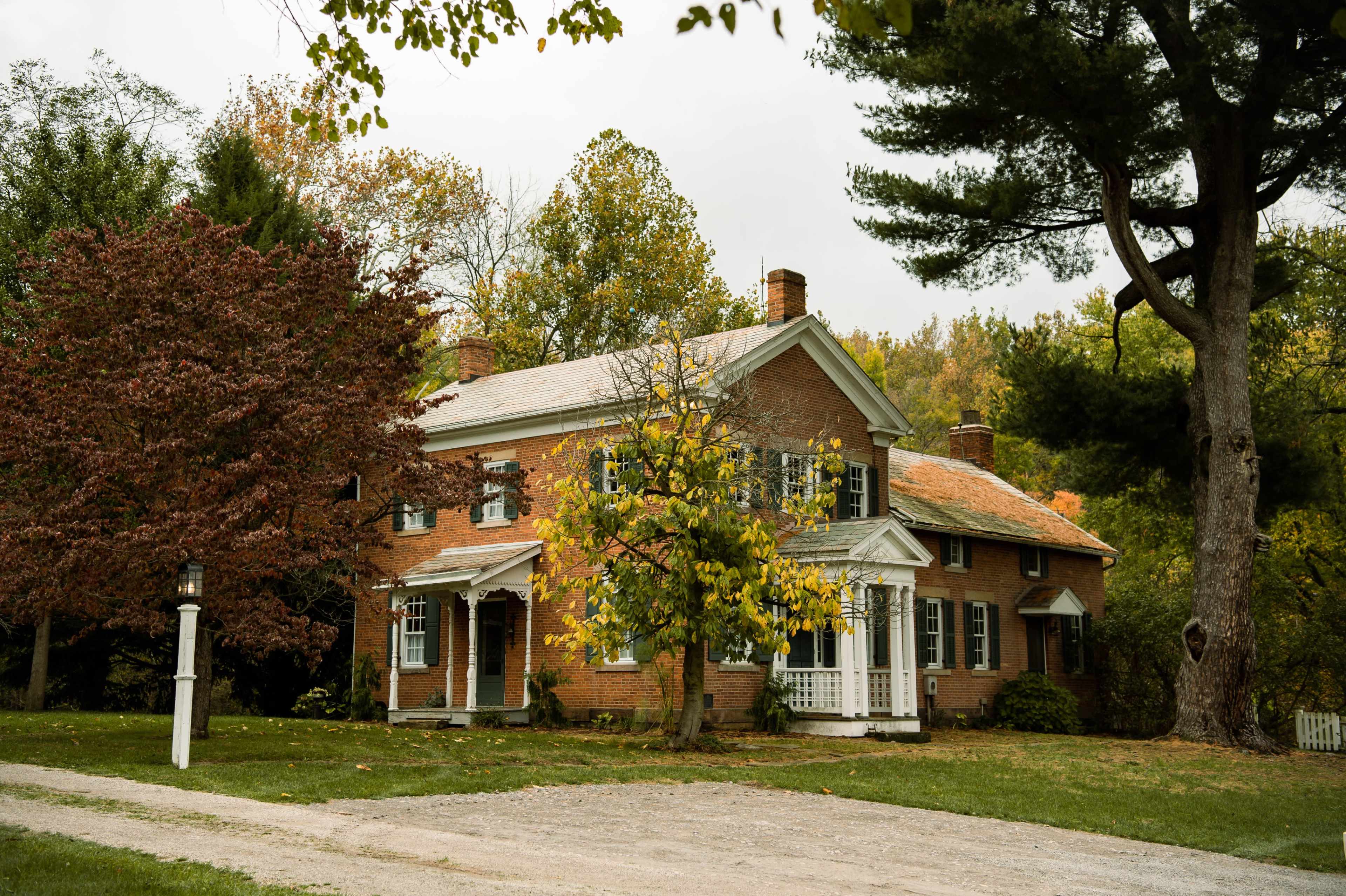 A two-story brick house with white trim is surrounded by trees and a gravel driveway.
