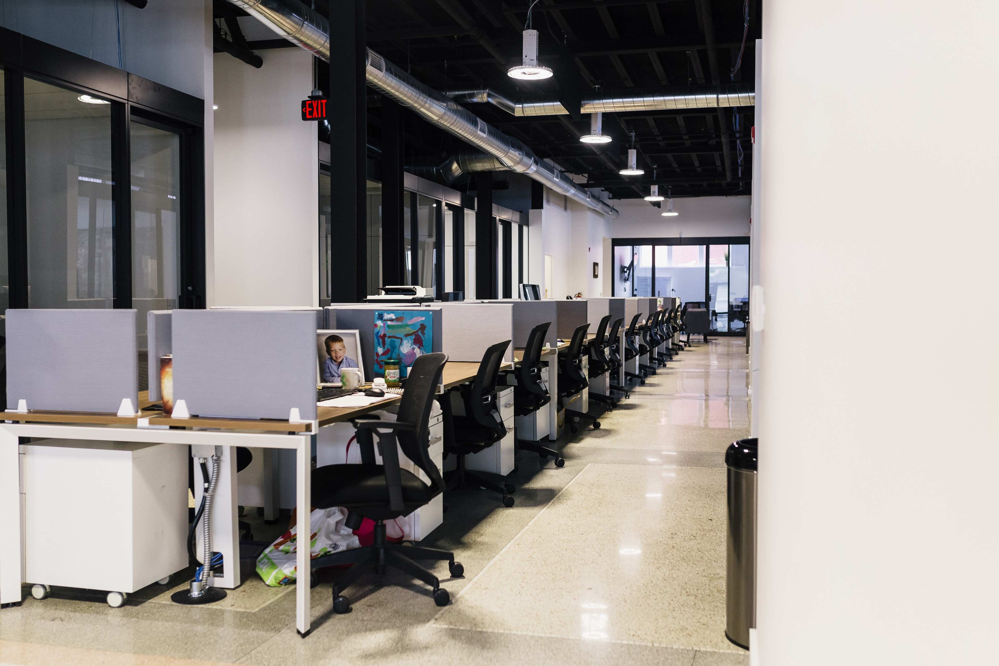 A row of empty office cubicles with computer monitors and chairs, alongside a polished floor and large windows.