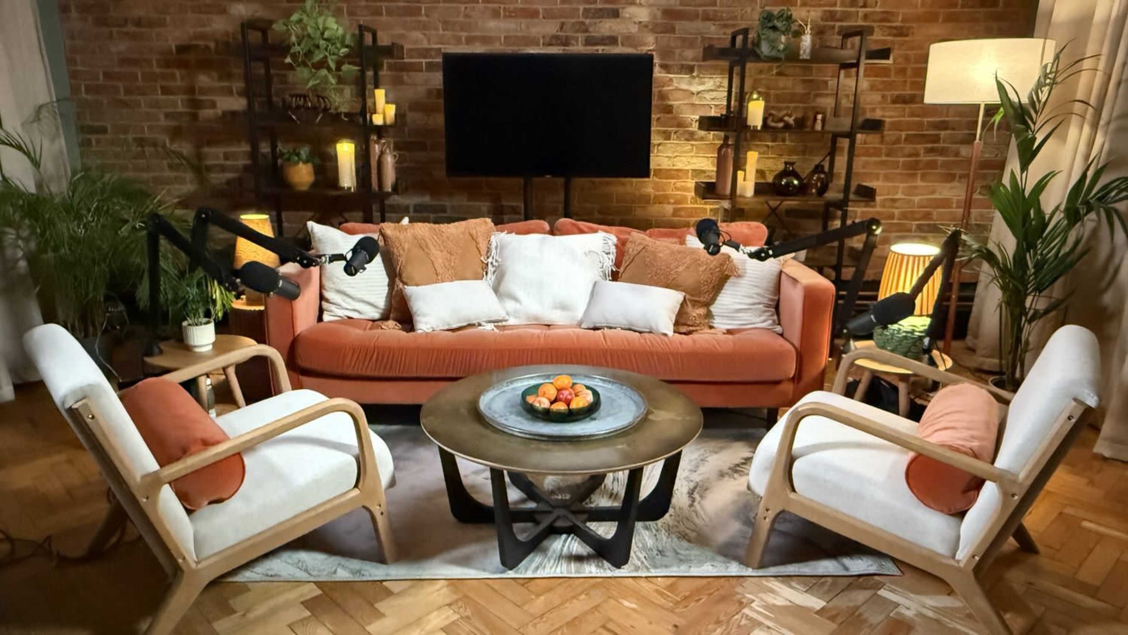 A cozy living room features an orange sofa with white and textured pillows, surrounded by two wooden chairs, a round coffee table with a bowl of oranges, and decorative plants against a brick wall.