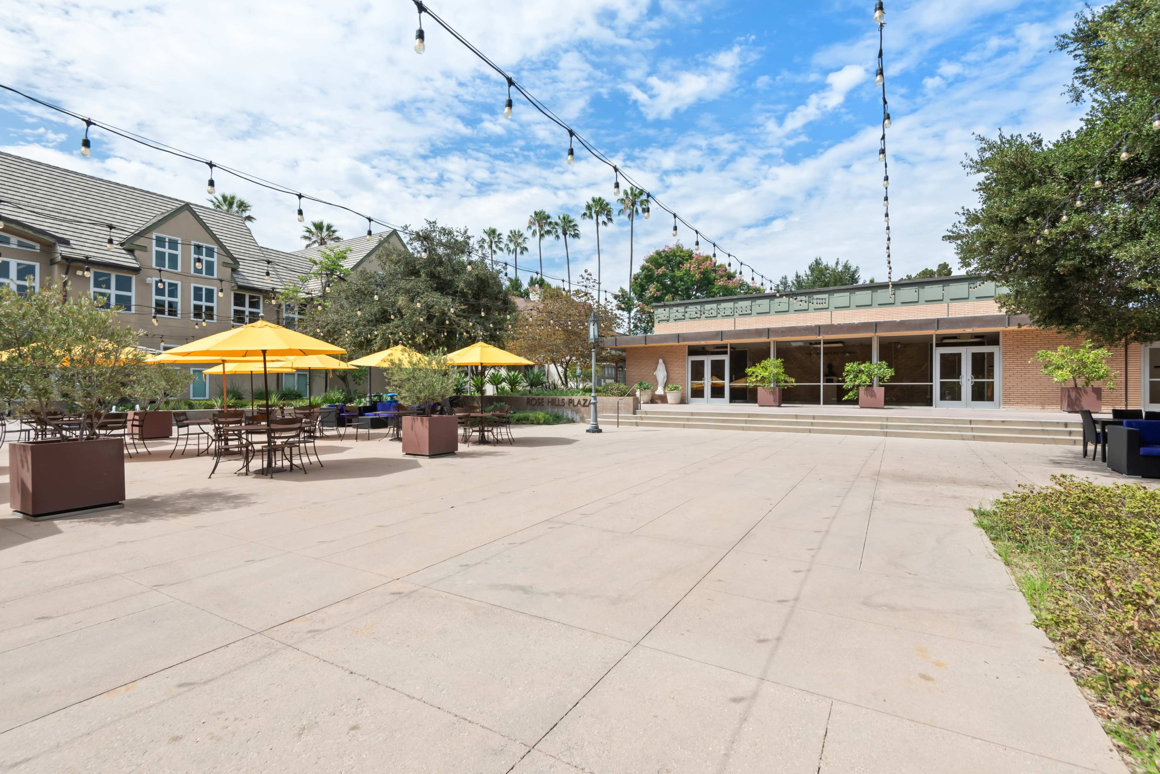The image shows an outdoor patio area with yellow umbrellas and tables, surrounded by greenery and buildings under a blue sky.