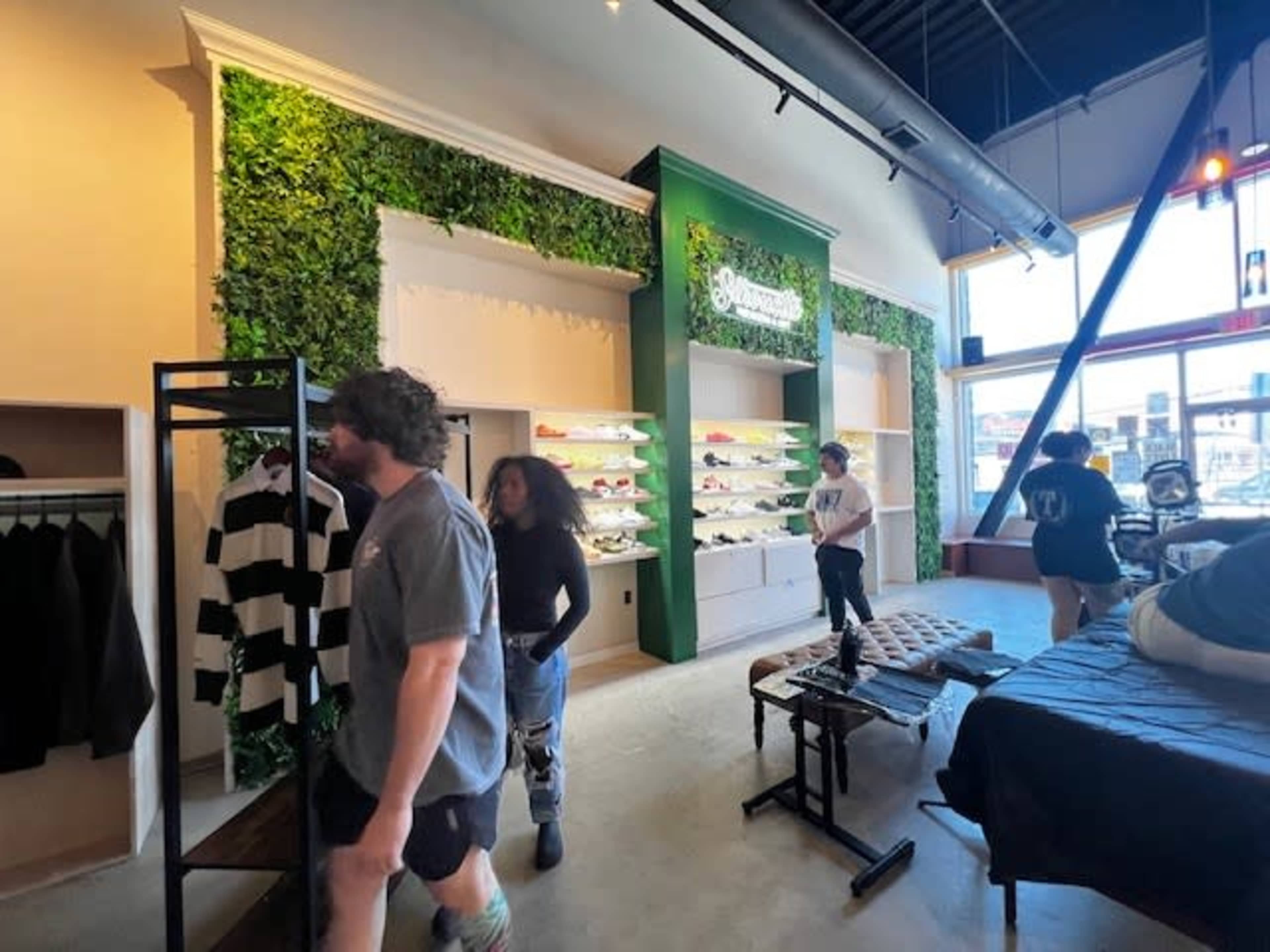 The image shows a clothing store interior with green foliage decor on the walls, clothing racks, and customers browsing the merchandise.