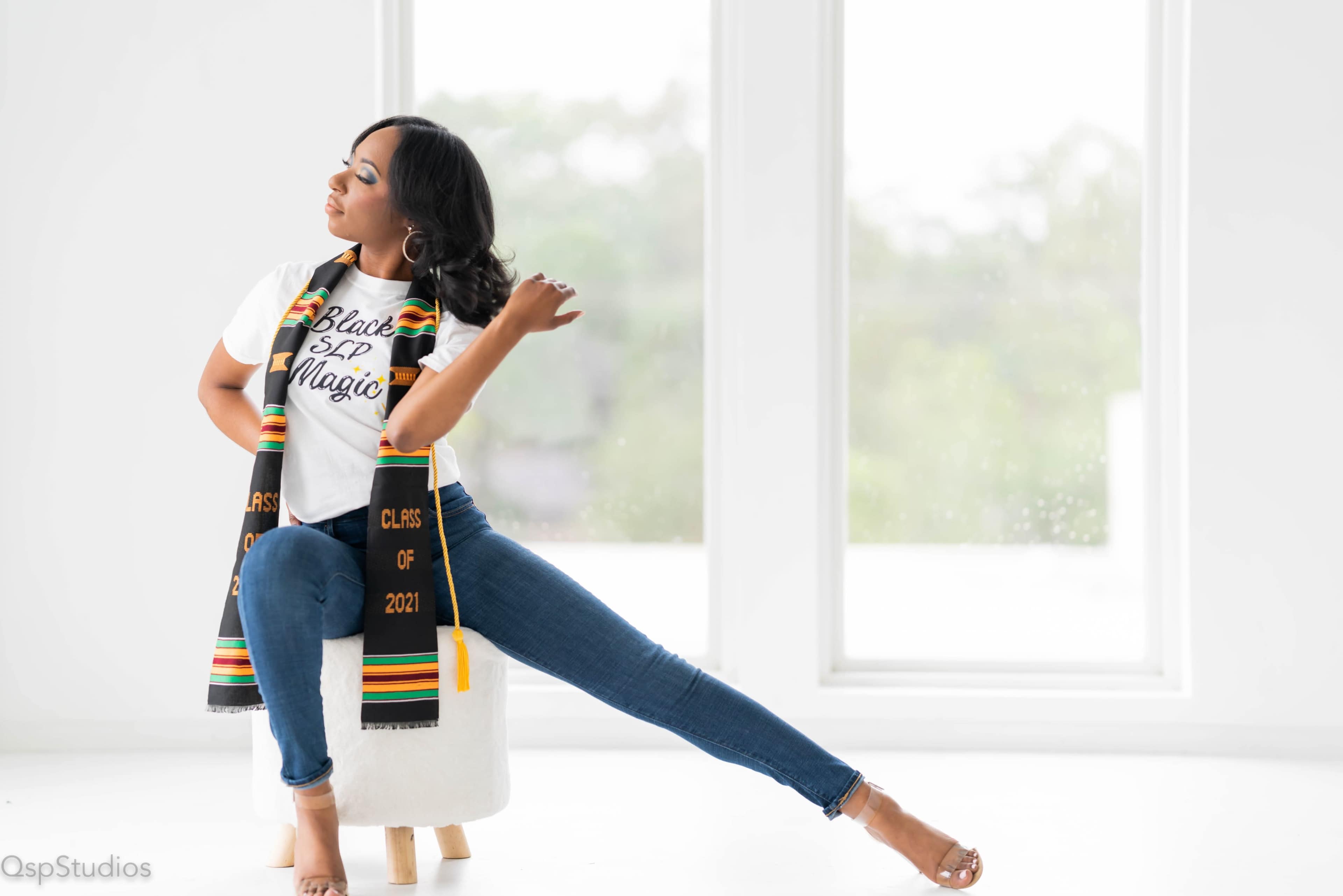 A young woman sits on a stool, wearing a "Black SIP Magic" t-shirt and a graduation scarf, while extending one leg out beside her.