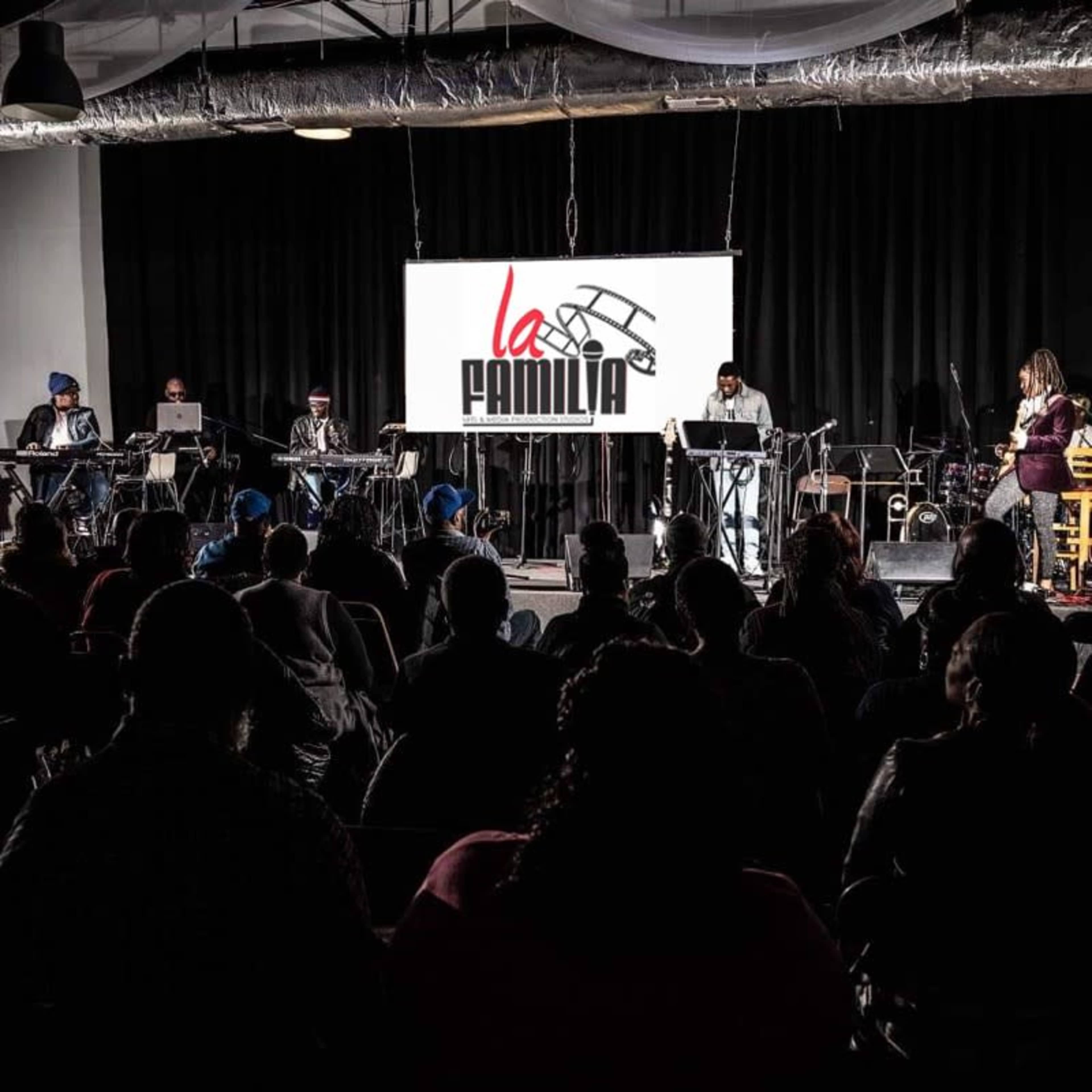 A band performs on stage in front of an audience, with a large screen displaying the logo "la FAMILIA" in the background.