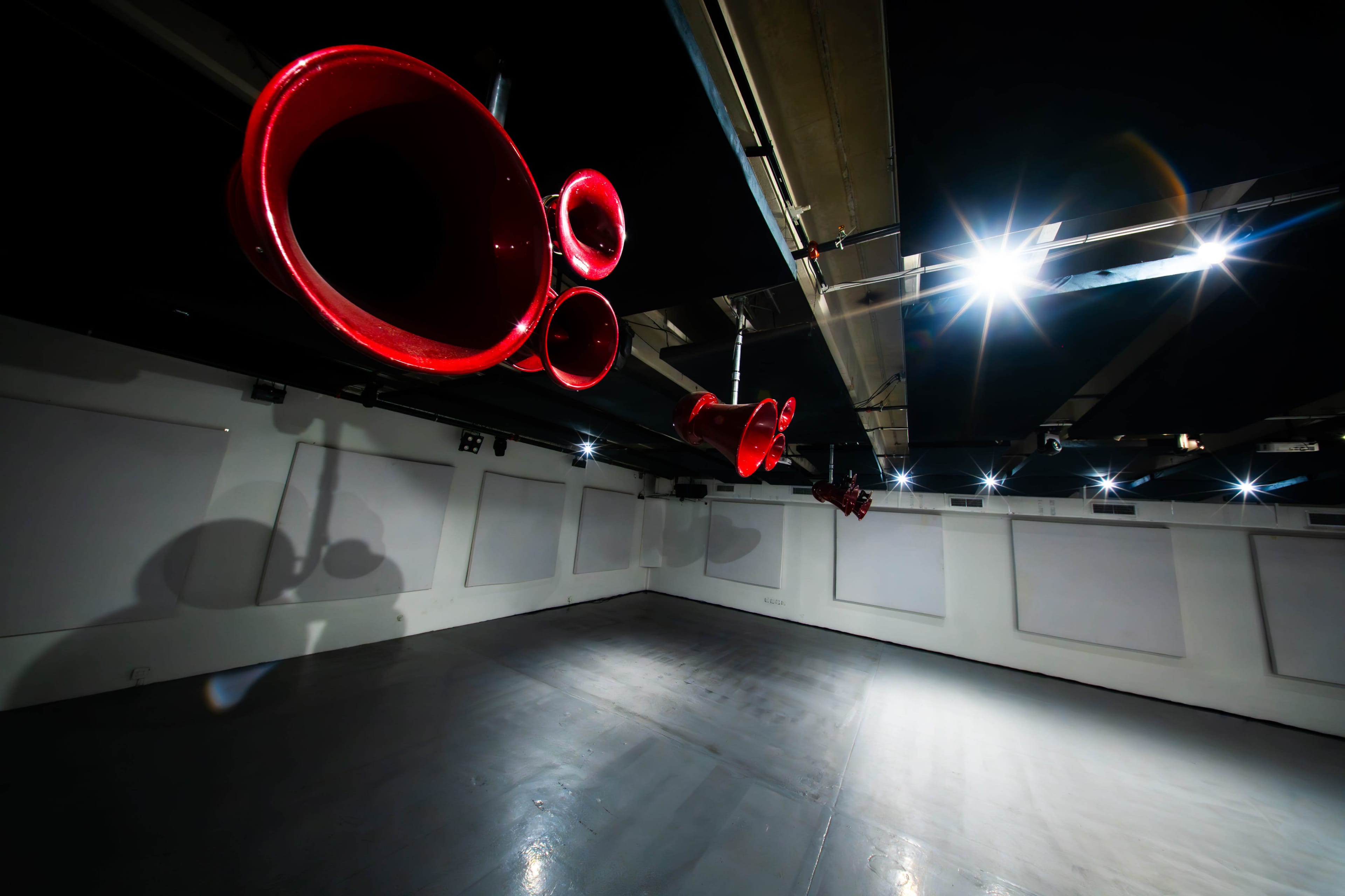 The image shows a dimly lit gallery or exhibition space with red circular sculptures suspended from the ceiling.