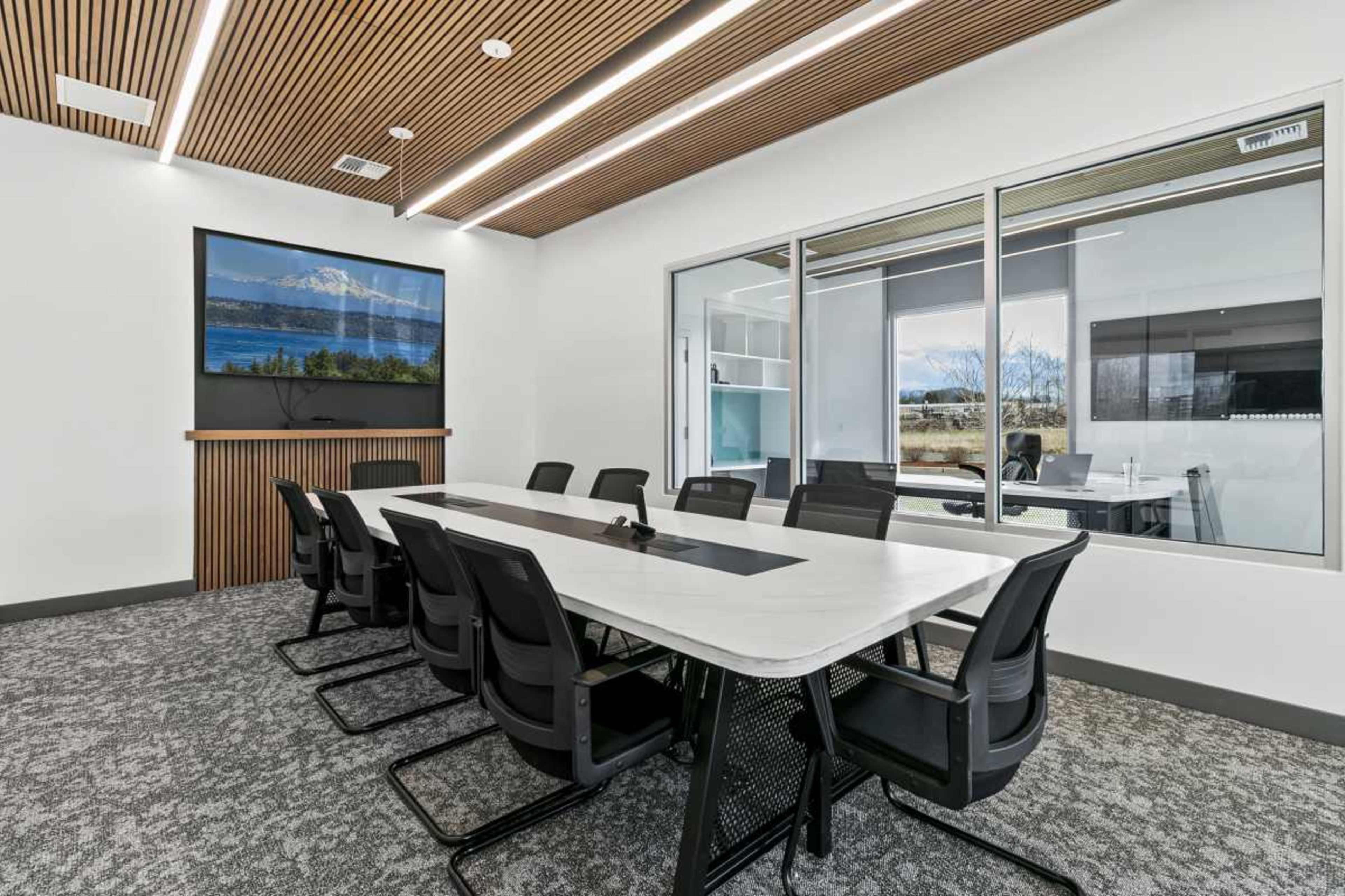 A modern conference room features a long white table surrounded by black chairs, with a large screen mounted on the wall and large windows offering a view outside.