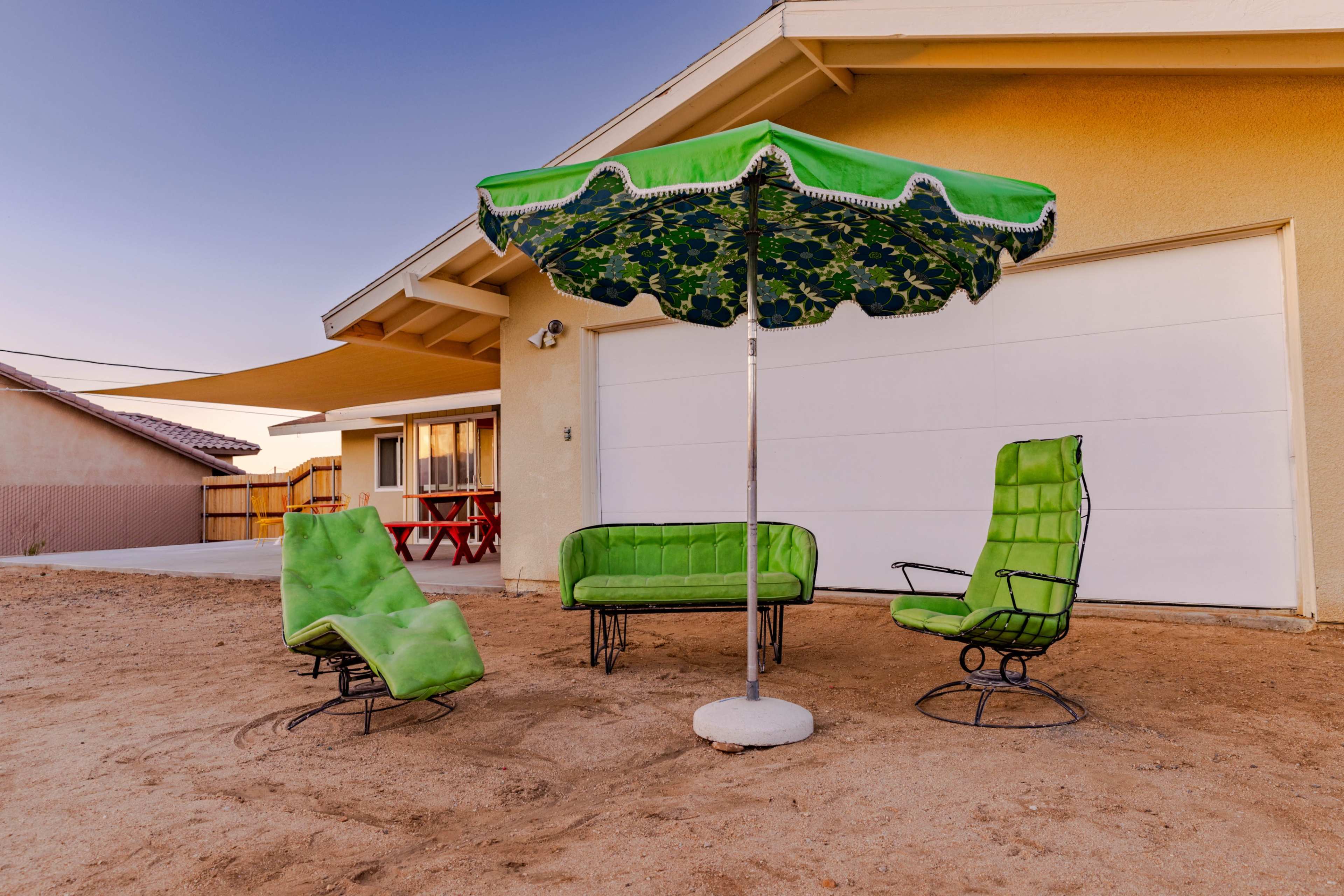 A set of green outdoor furniture, including two chairs, a sofa, and a large umbrella, is arranged on a sandy surface next to a garage.