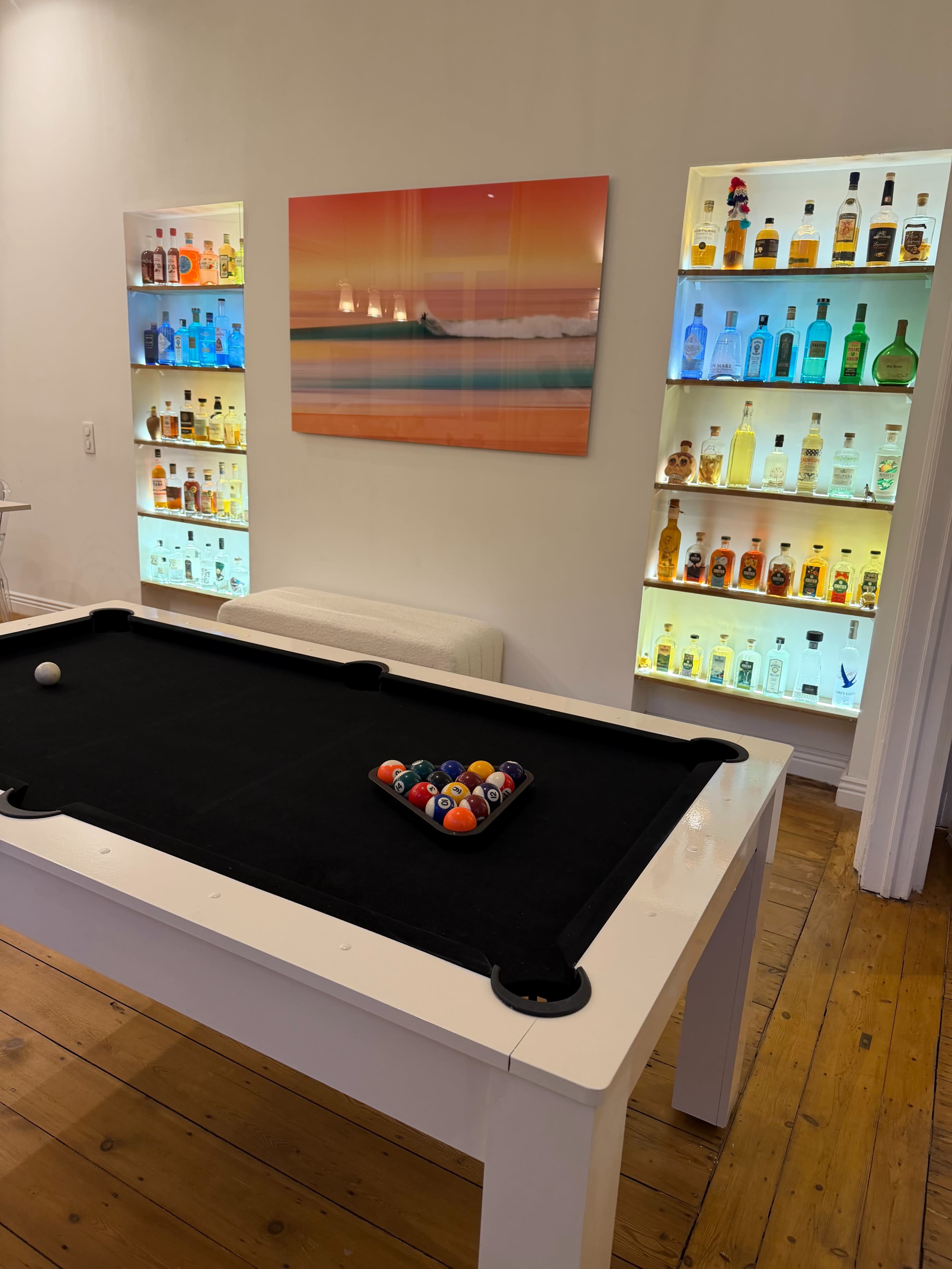 The image shows a pool table with a triangle of balls arranged on it, surrounded by illuminated shelves displaying various bottles of liquor and a colorful wall art piece featuring a beach scene.