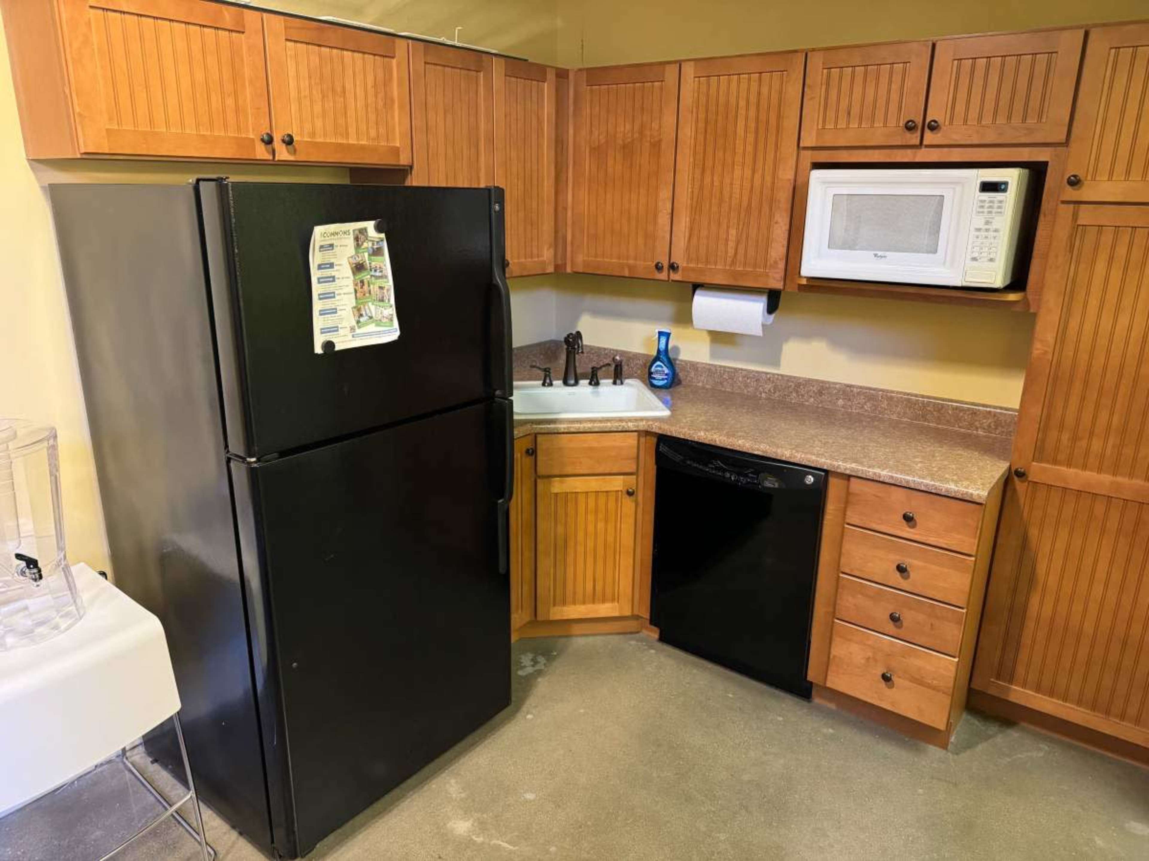 The image shows a kitchen with wooden cabinets, a black refrigerator, a black dishwasher, and a countertop with a microwave and sink.