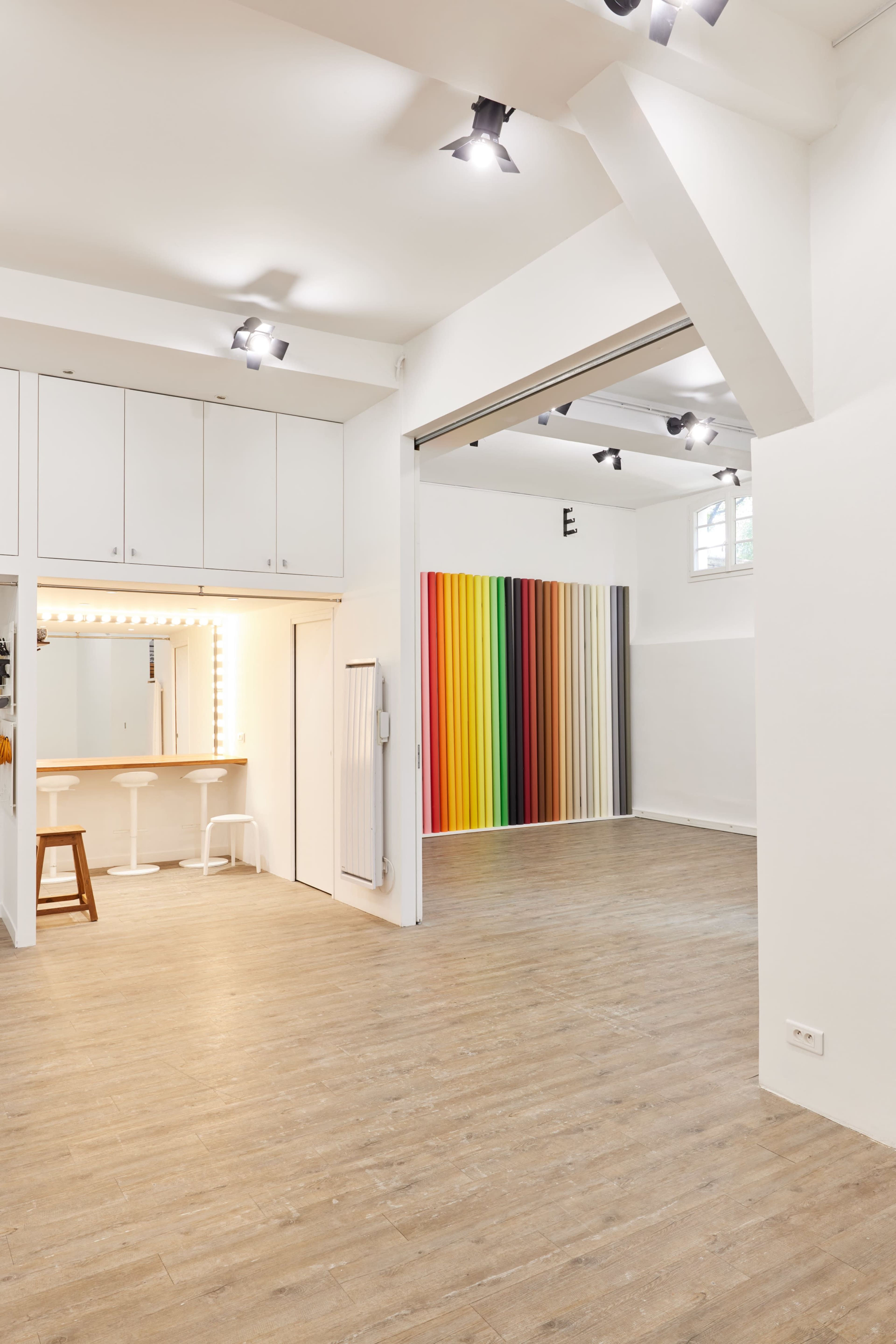 The image shows a spacious studio with light wooden flooring, featuring a bar area with two stools and a wall of colorful vertical stripes.