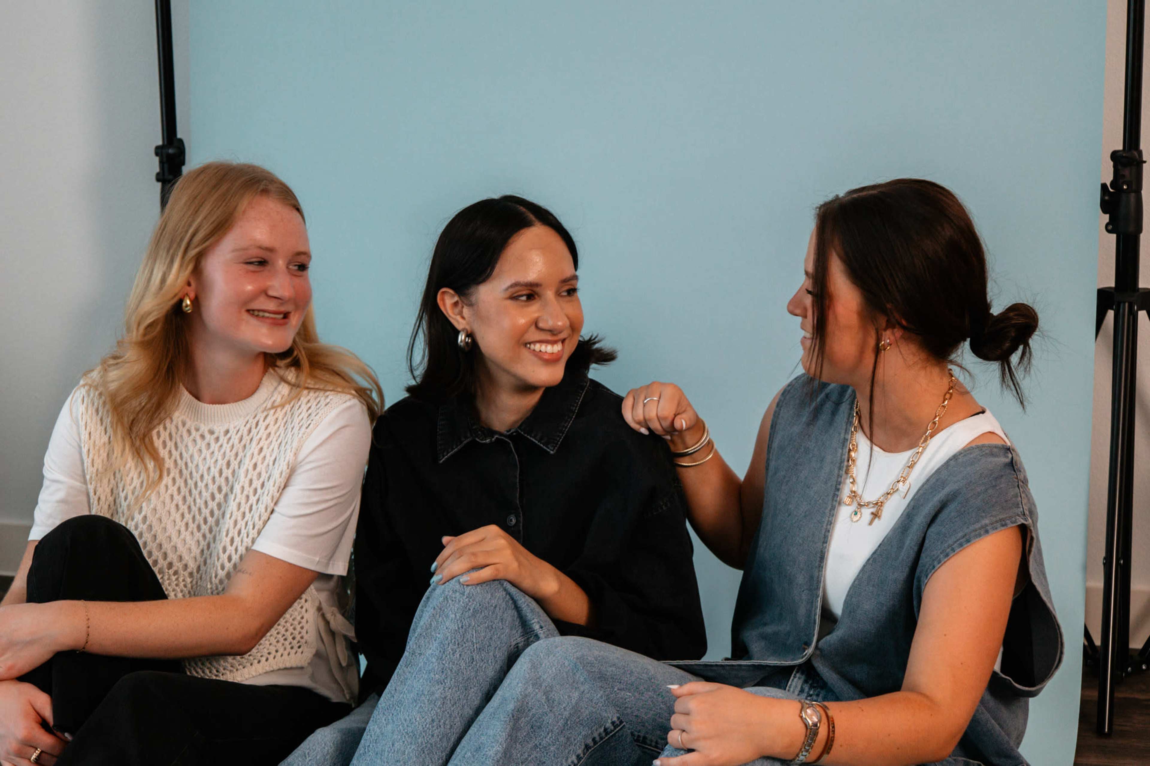 Three young women sit on a floor in front of a light blue backdrop, engaged in conversation and smiling at each other.