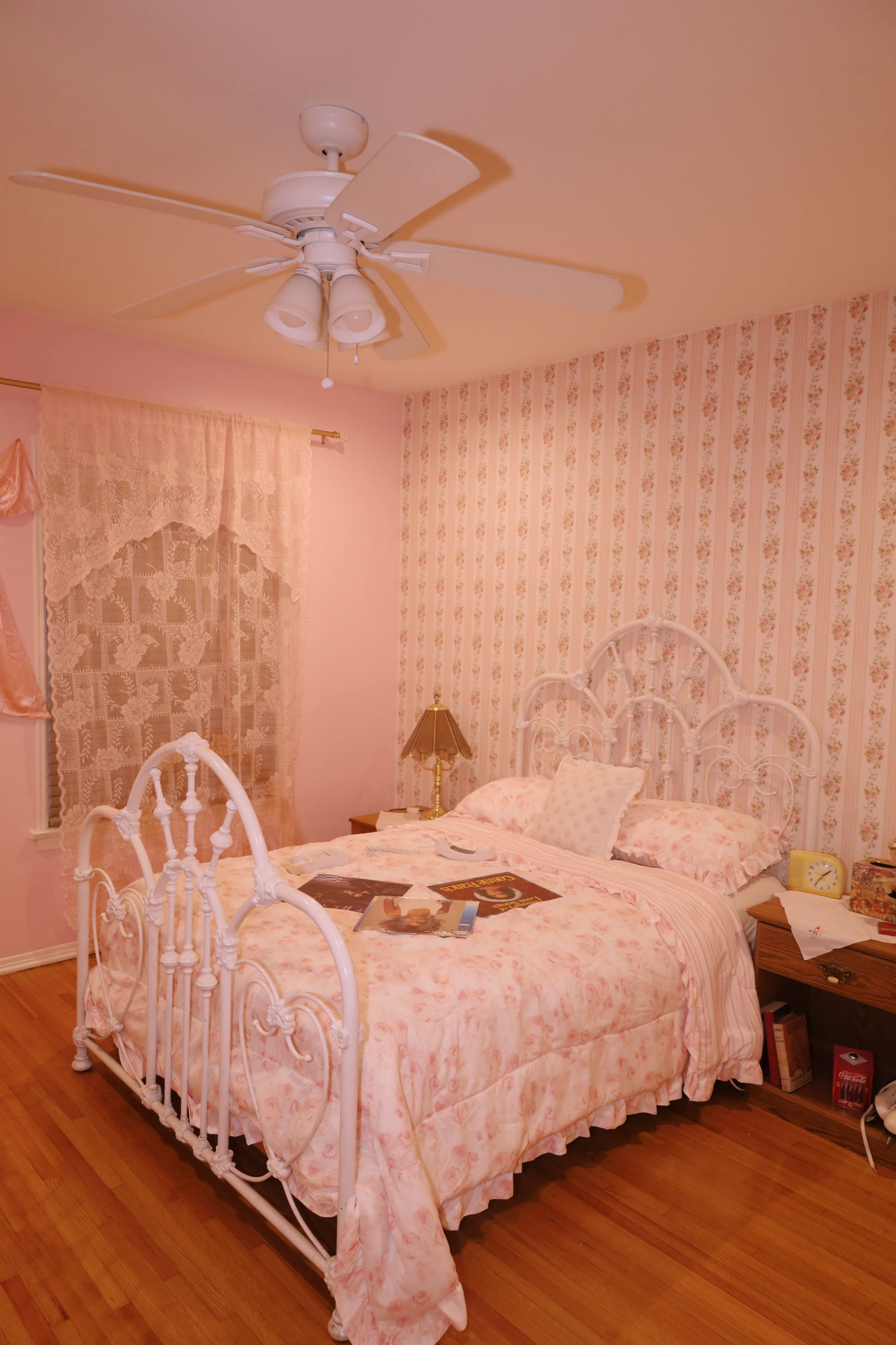 The image shows a pink-themed bedroom featuring a white metal bed with floral bedding, a bedside table, and patterned wallpaper.
