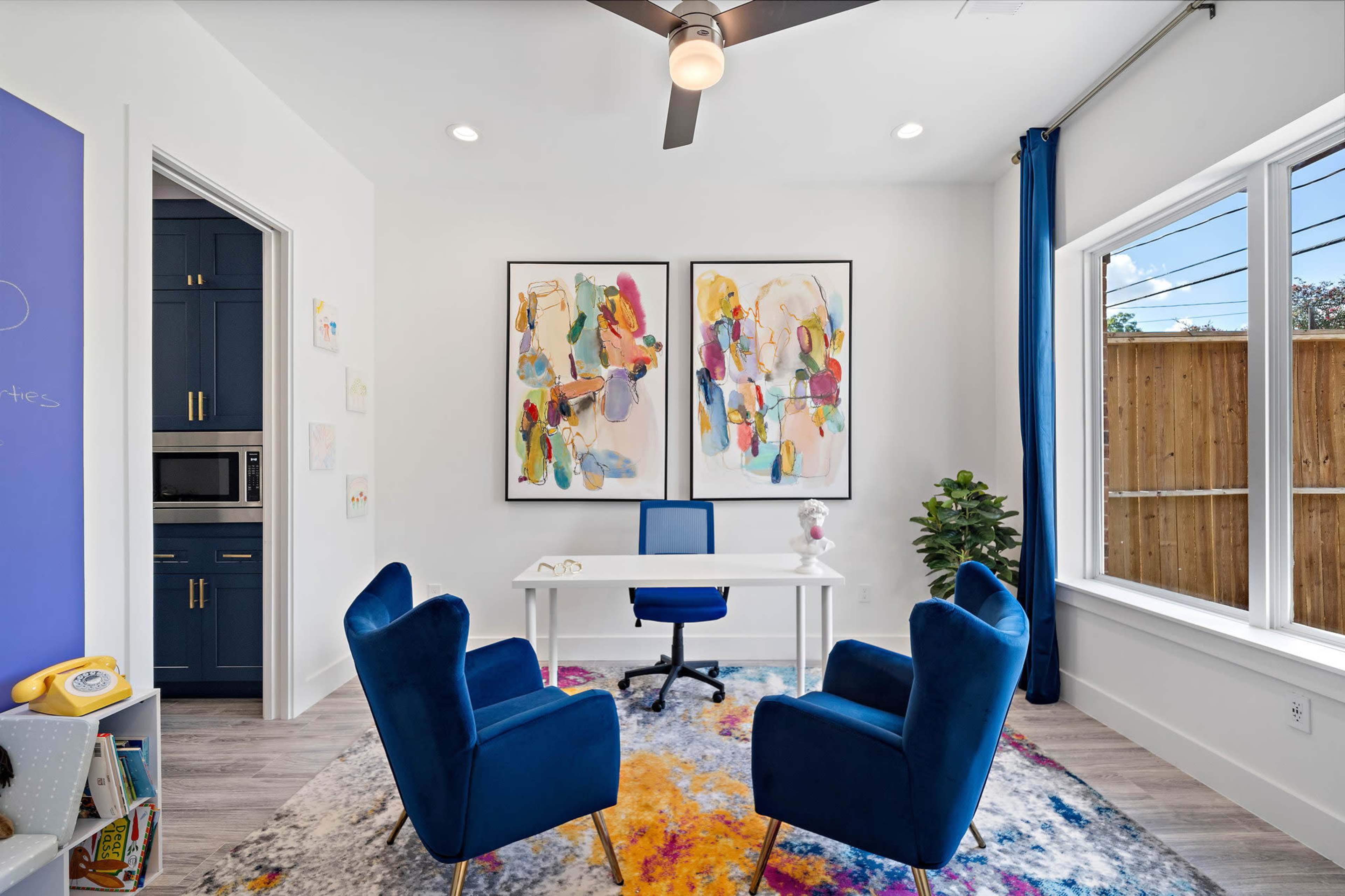 A bright home office features two blue chairs, a white desk, colorful wall art, and large windows with a view of a wooden fence.