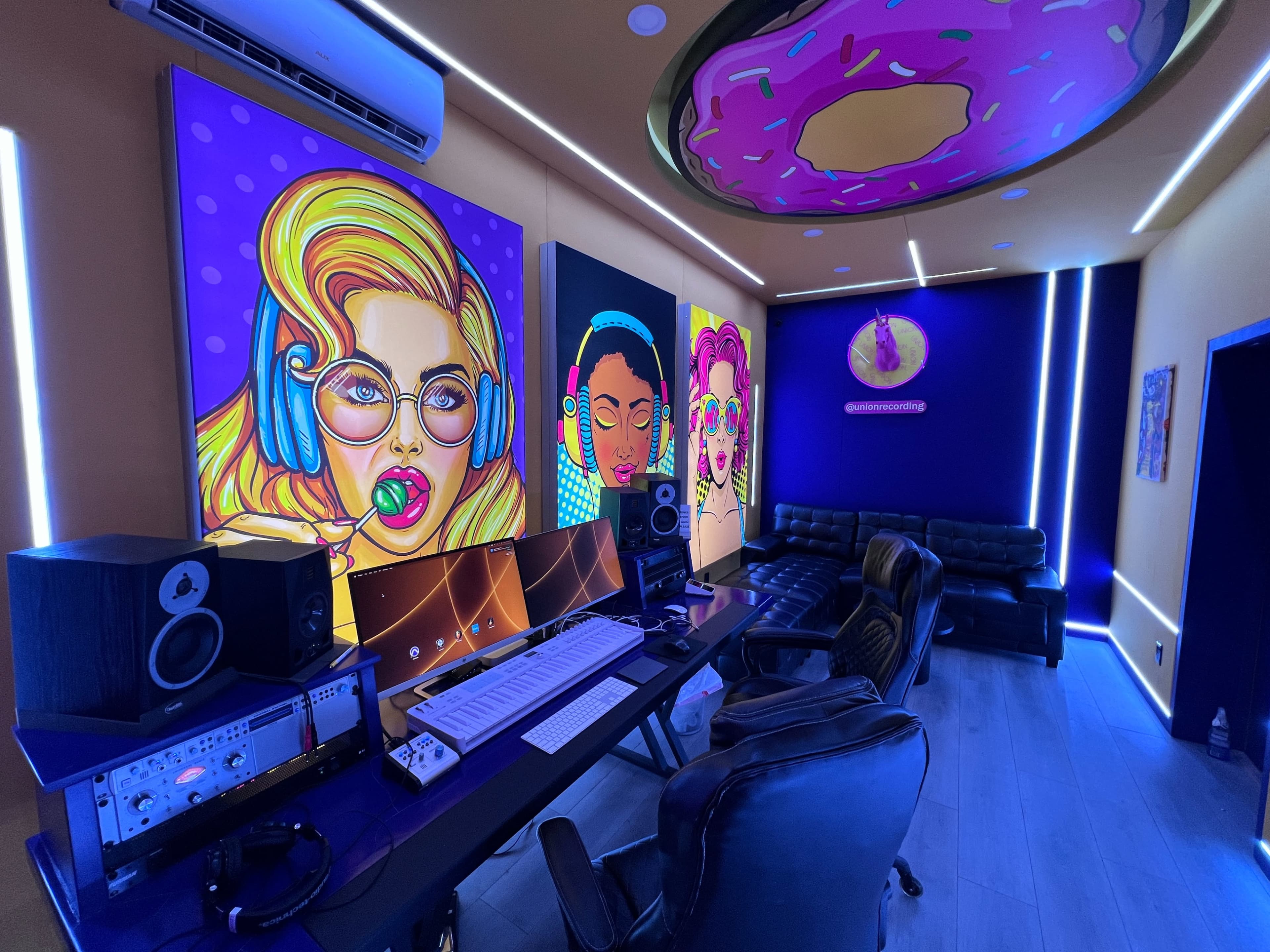 The image shows a vibrant recording studio featuring colorful artwork, a modern desk with audio equipment, and a black seating area.