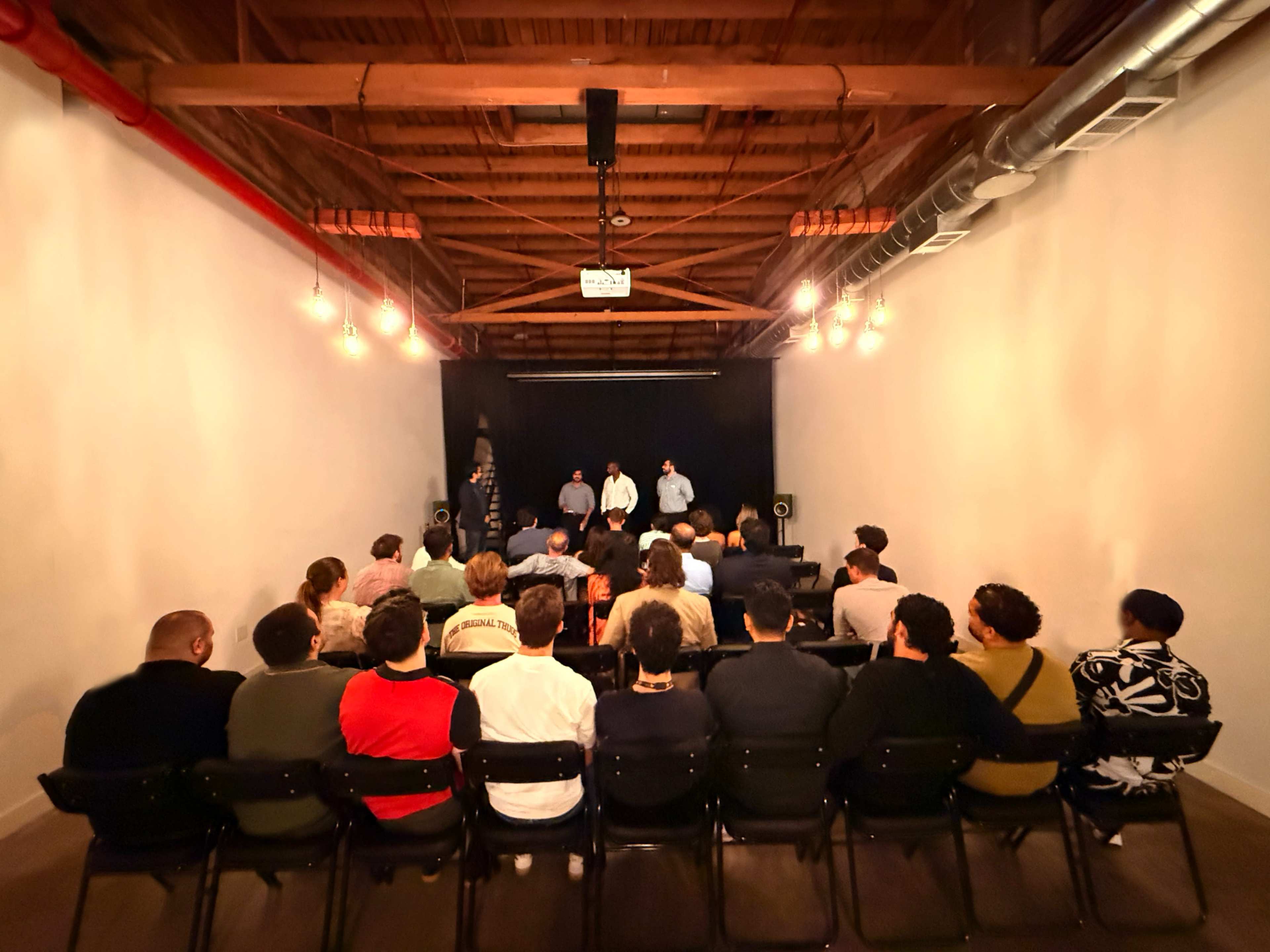 A group of people sits in chairs facing a small stage where two individuals are speaking, with warm lighting illuminating the scene.