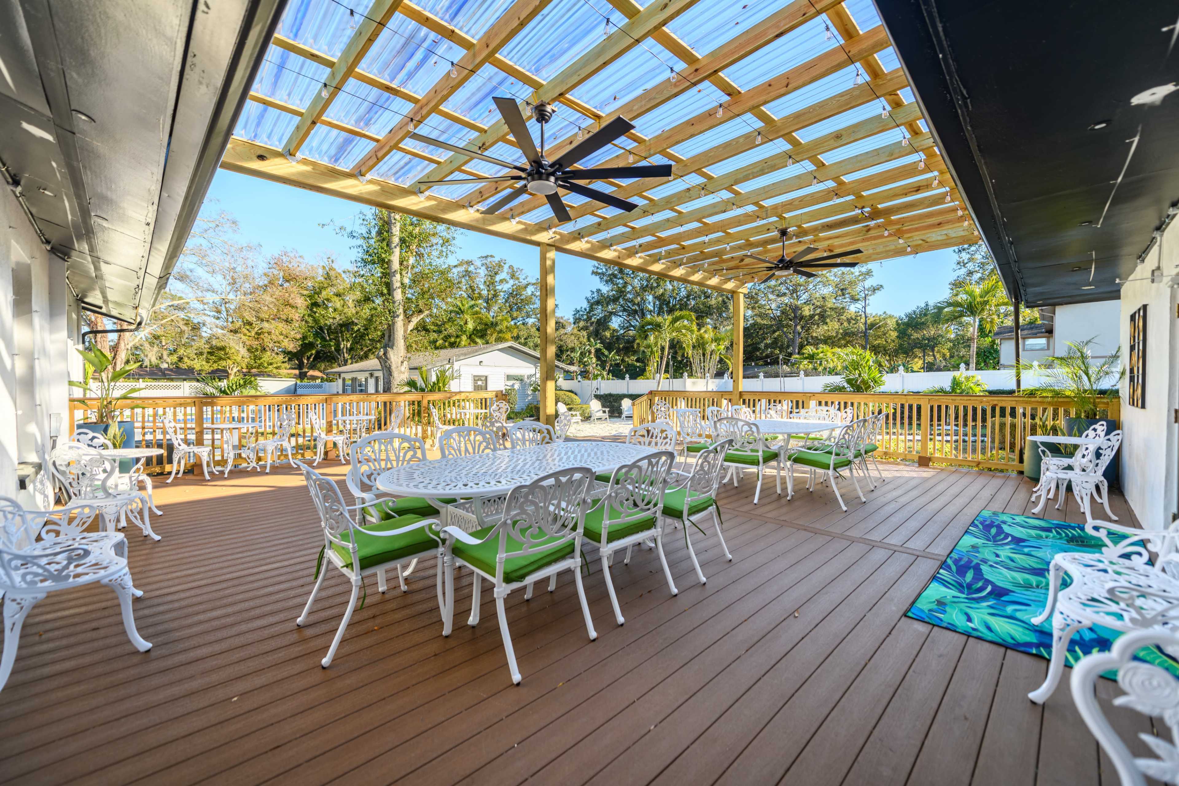 The image shows a spacious outdoor deck with white metal tables and chairs, featuring a wooden canopy overhead and surrounded by greenery.