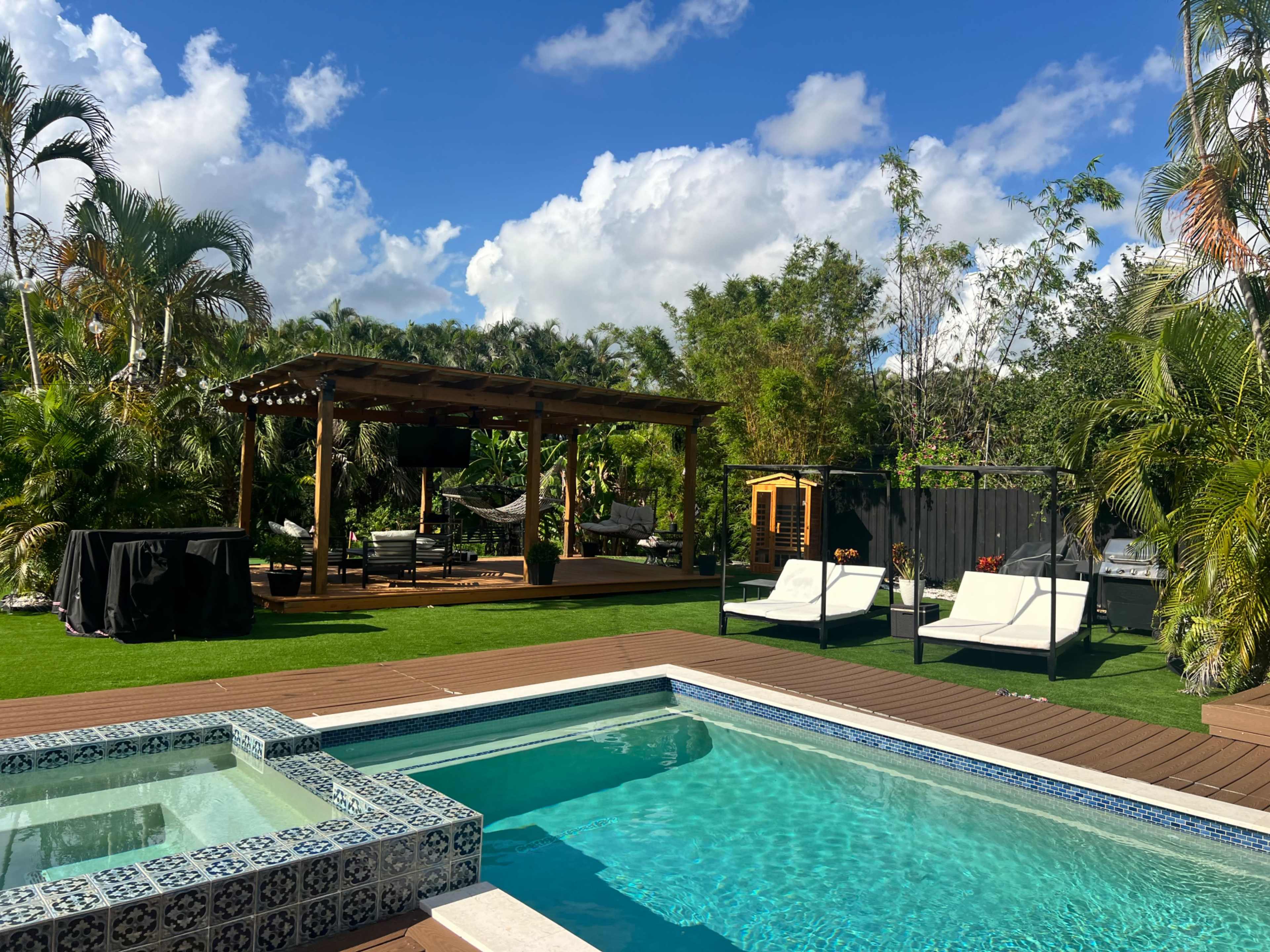 The image depicts a backyard oasis featuring a swimming pool with a spa, lounge chairs, a wooden pergola with seating, and tropical greenery under a blue sky.