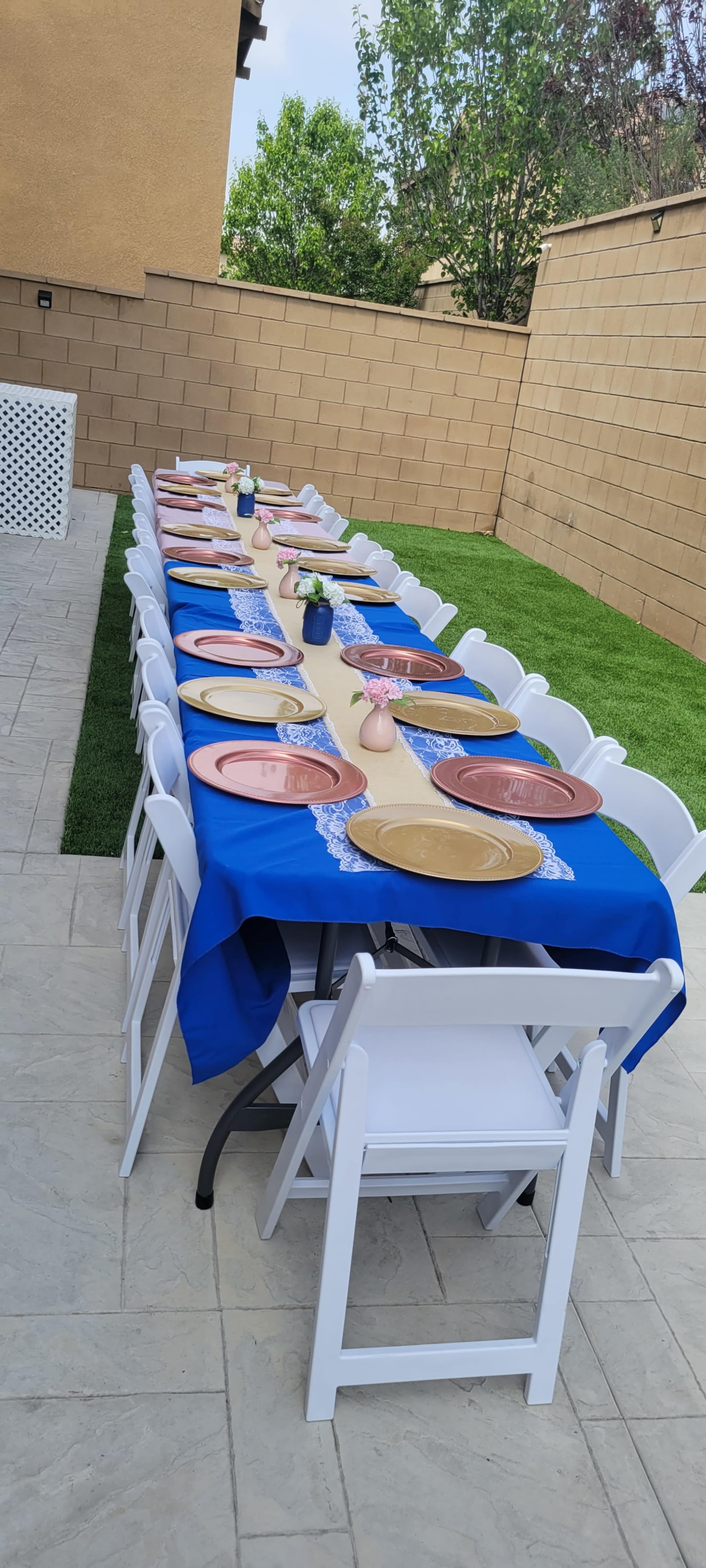 A long banquet table is set outdoors with blue tablecloths and arranged plates along its length, surrounded by white chairs on a grassy area.