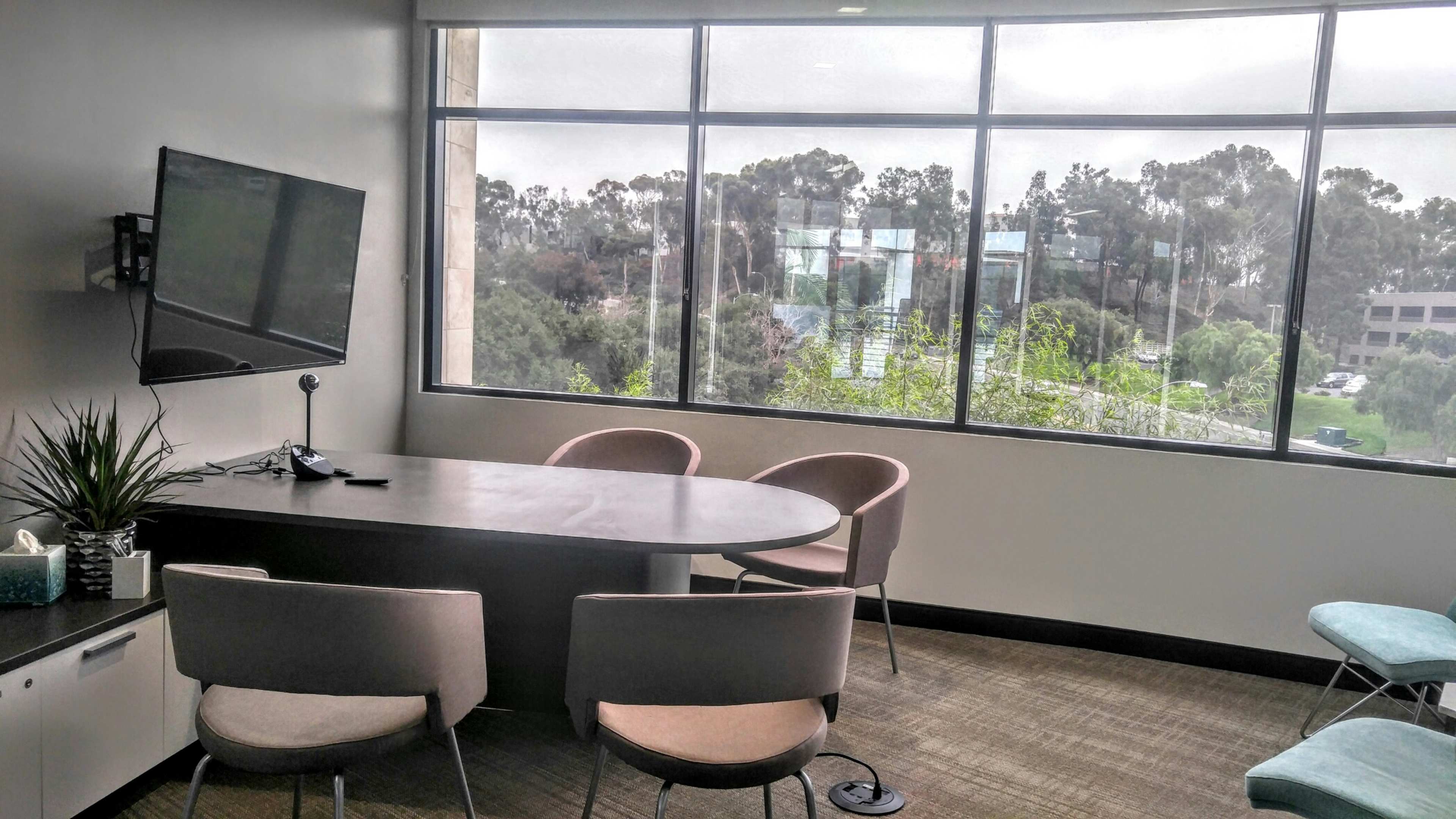 The image shows a conference room with a round table, four chairs, a wall-mounted television, and large windows overlooking a treed landscape.