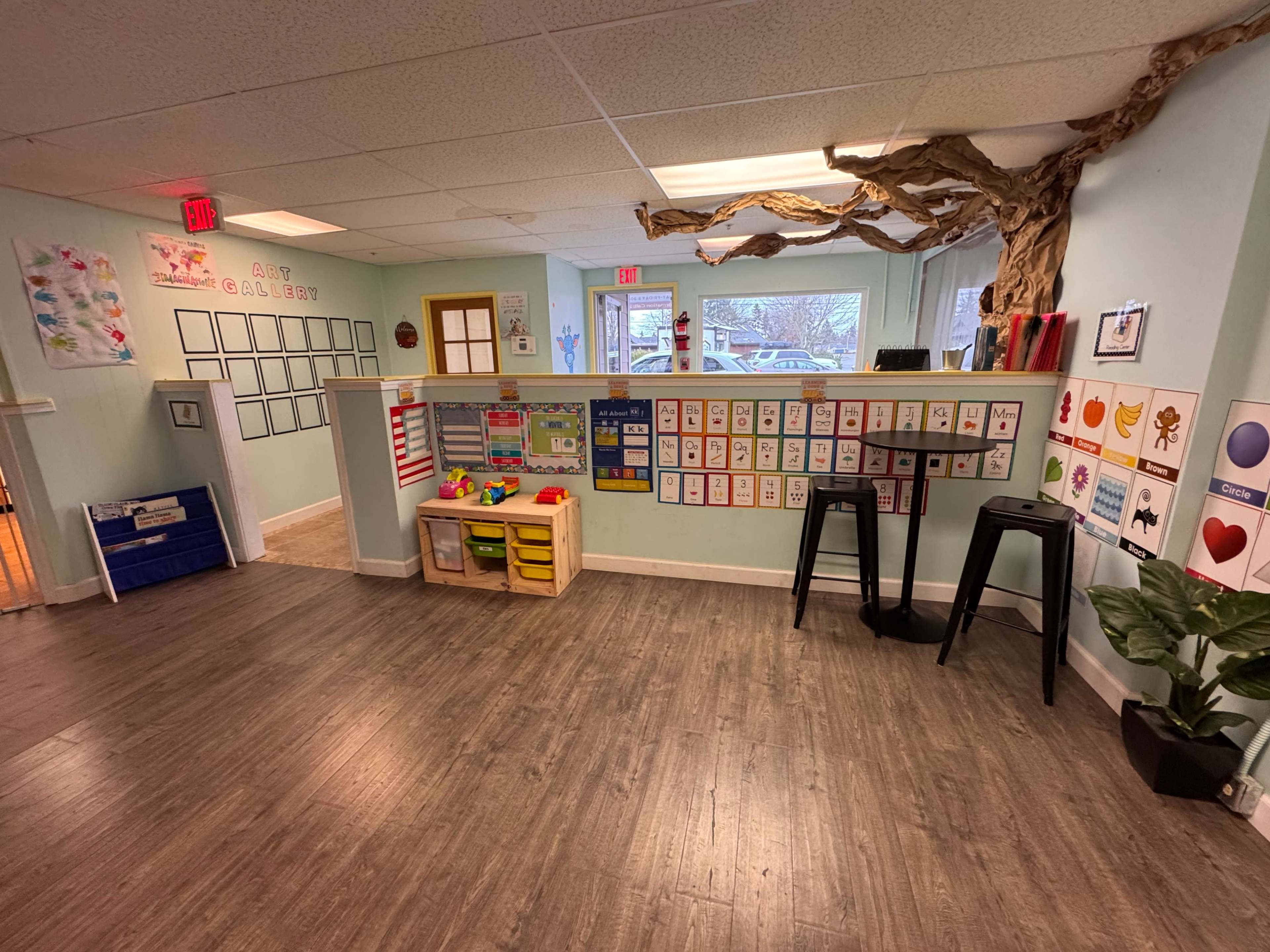 The image shows a classroom with colorful educational posters on the walls, a play area with toys, and wooden flooring.