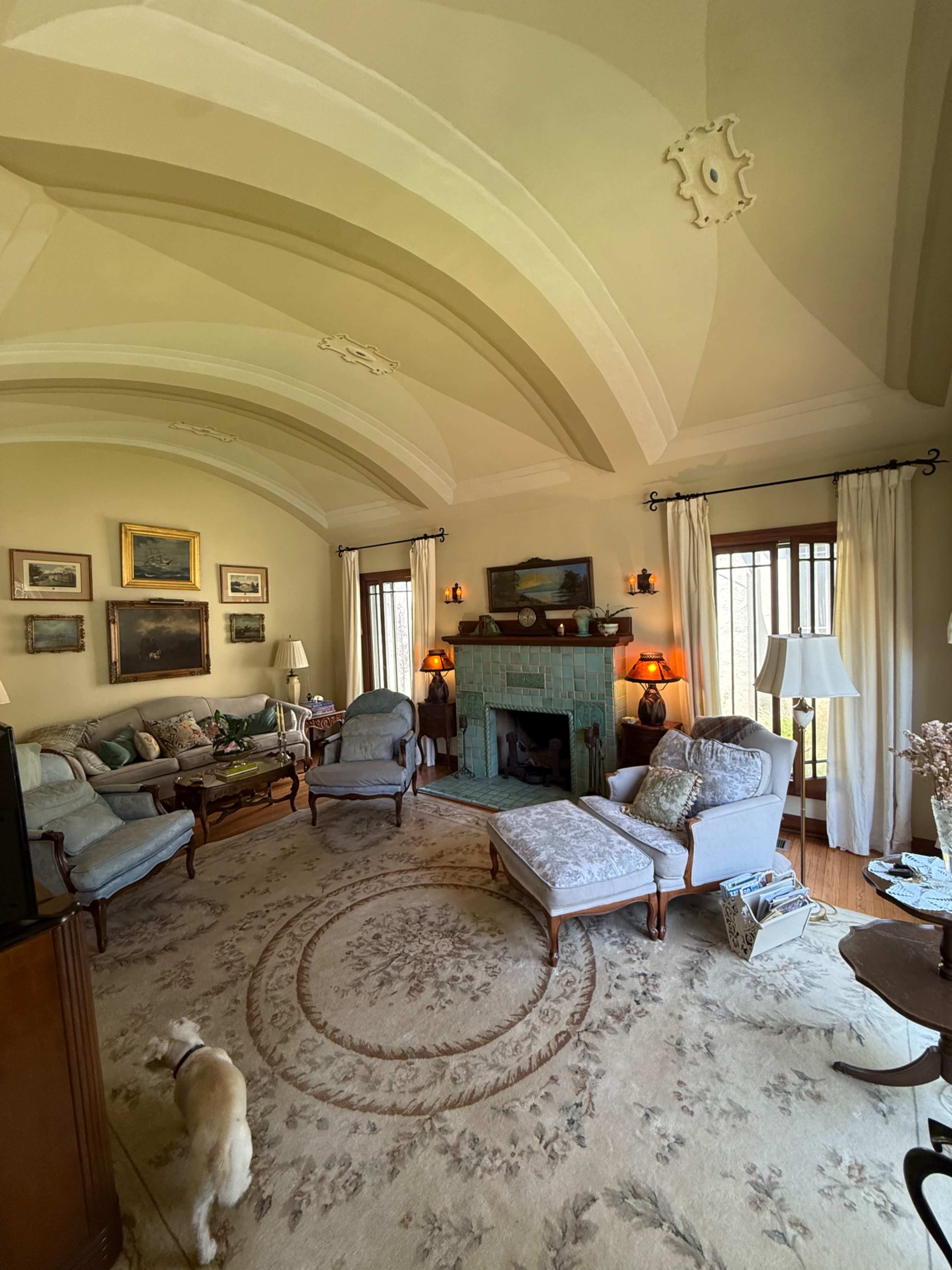 The living room features a high, arched ceiling, a turquoise fireplace, and several pieces of artwork on the walls, with a dog walking on a patterned rug.
