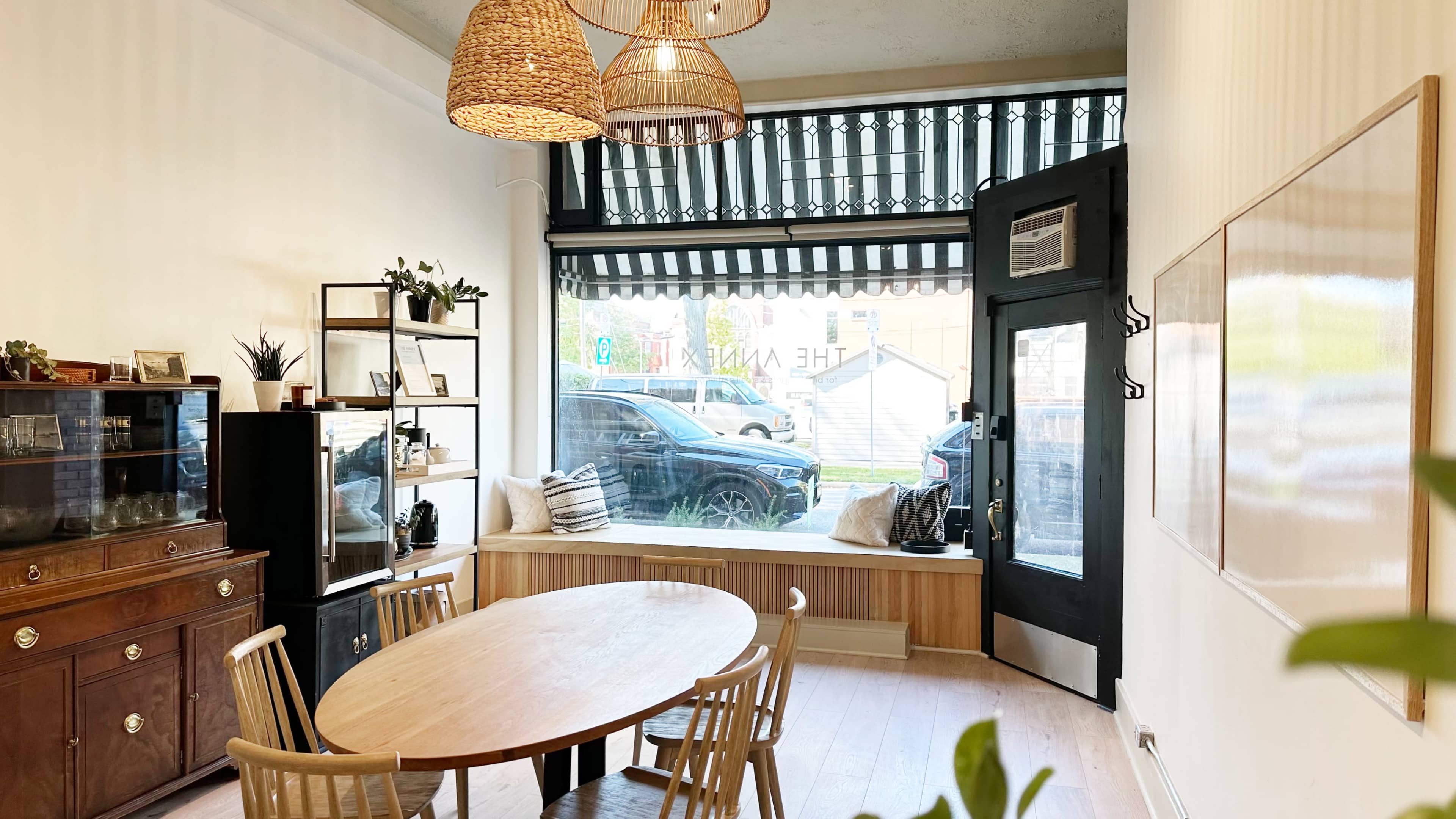 A well-lit room with a wooden table and chairs, a window seat, and a view of a street outside, featuring decorative lighting and shelving units.