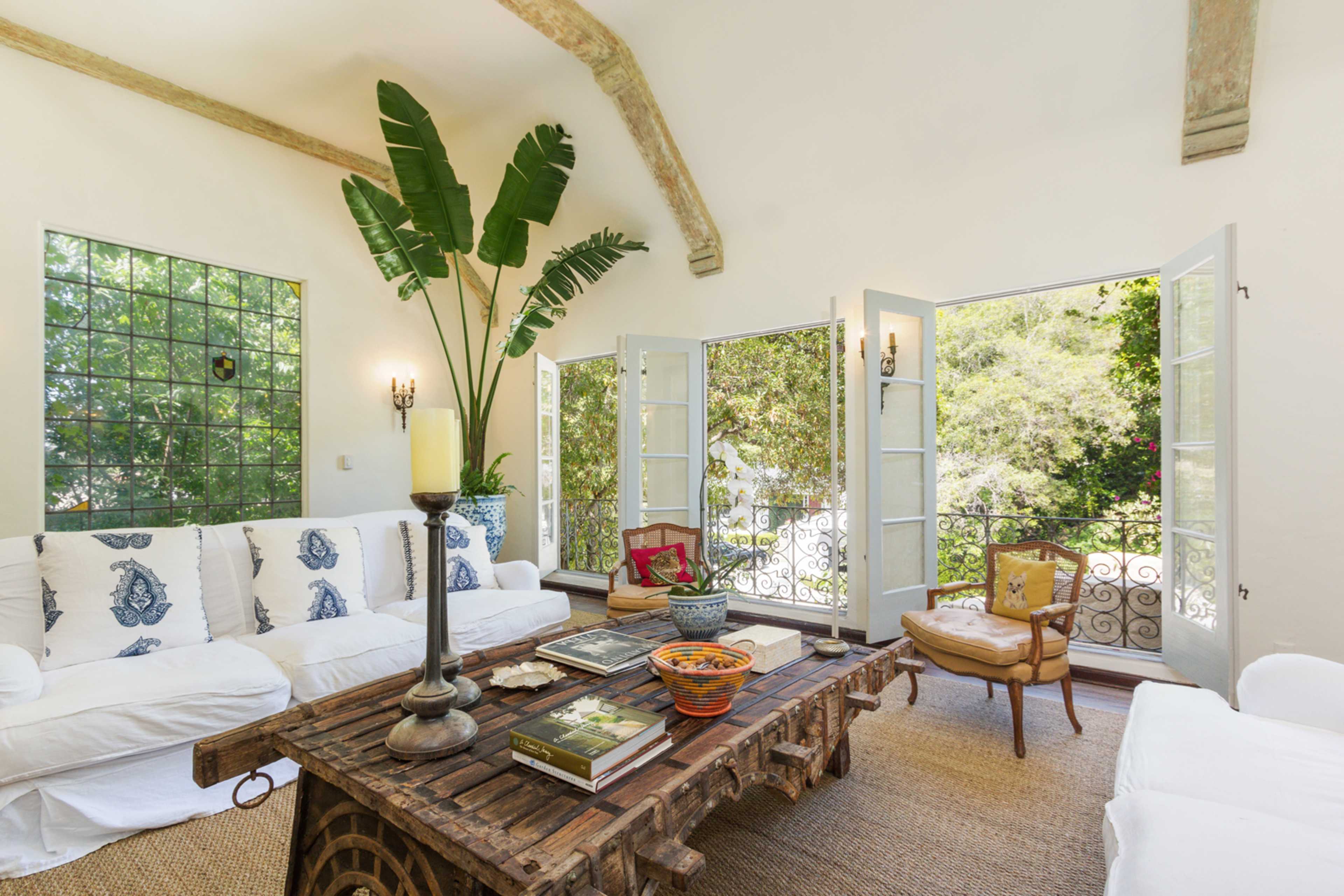 The scene shows a bright living room with white furniture, large windows opening to a view of greenery, and a rustic wooden coffee table adorned with books and decorative items.