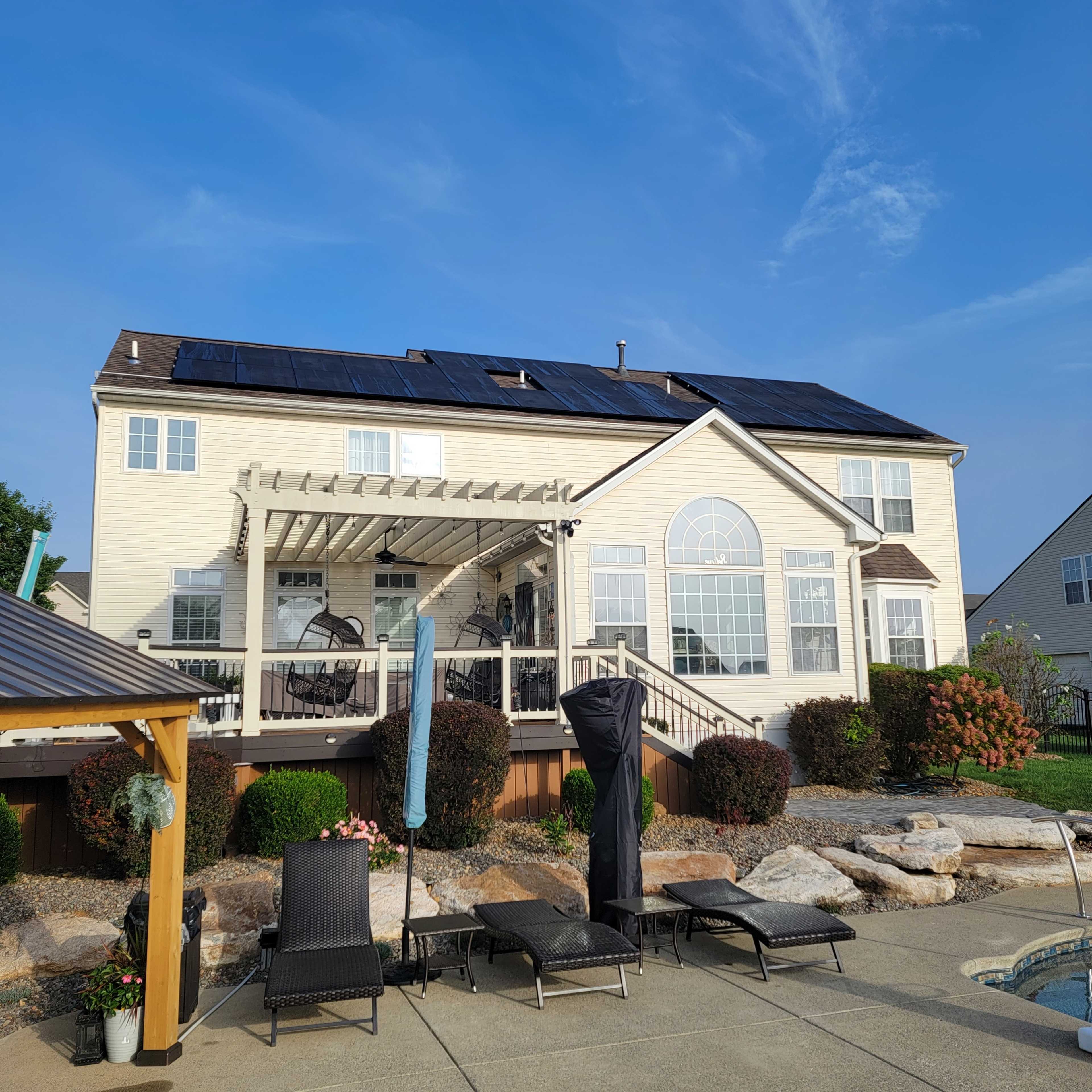 A two-story house with solar panels on the roof, backed by a spacious patio and a swimming pool area featuring lounge chairs and landscaping.