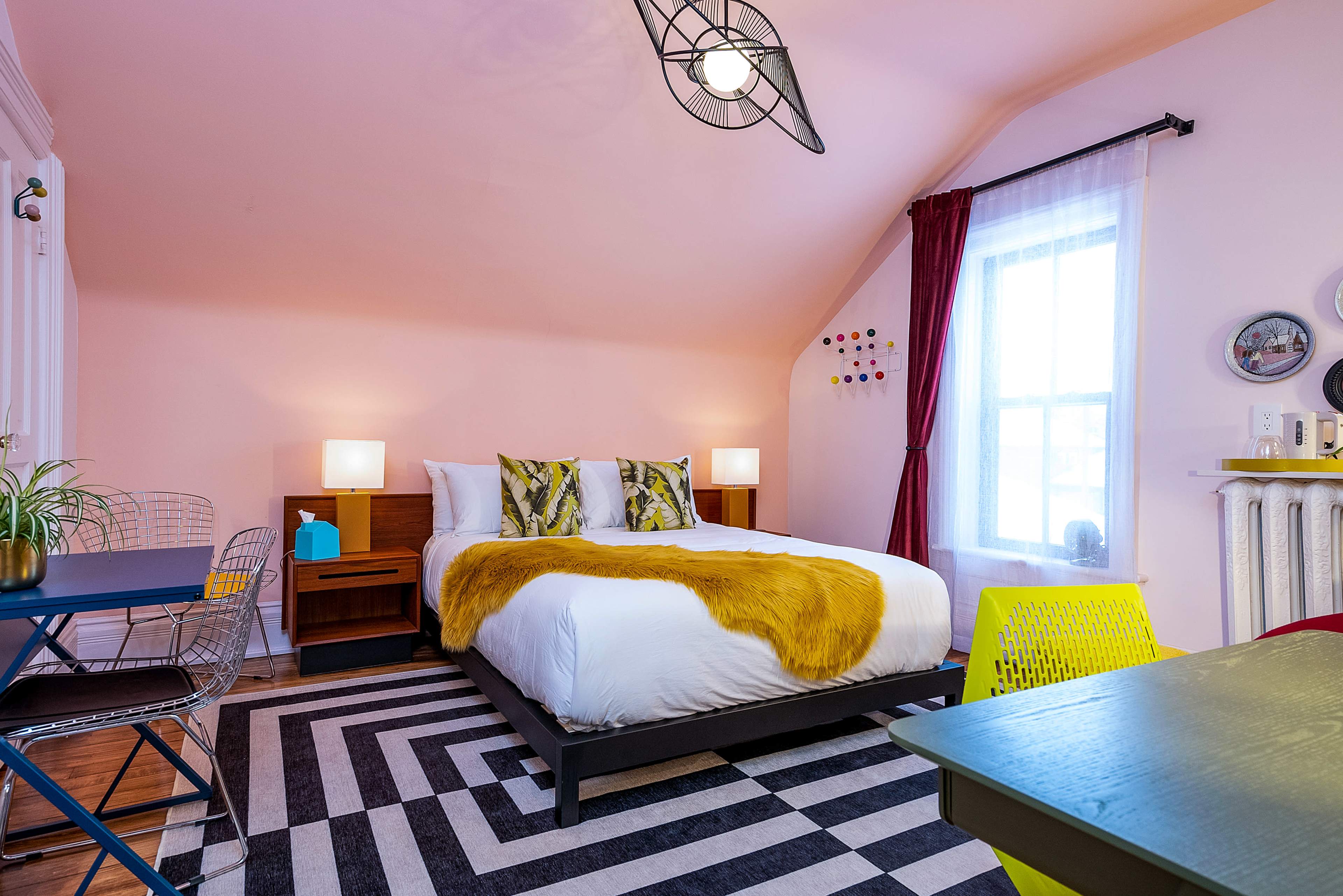 A cozy bedroom with a large bed dressed in white linens and a yellow throw, complemented by a geometric black and white rug, a small dining area, and a window with sheer curtains.