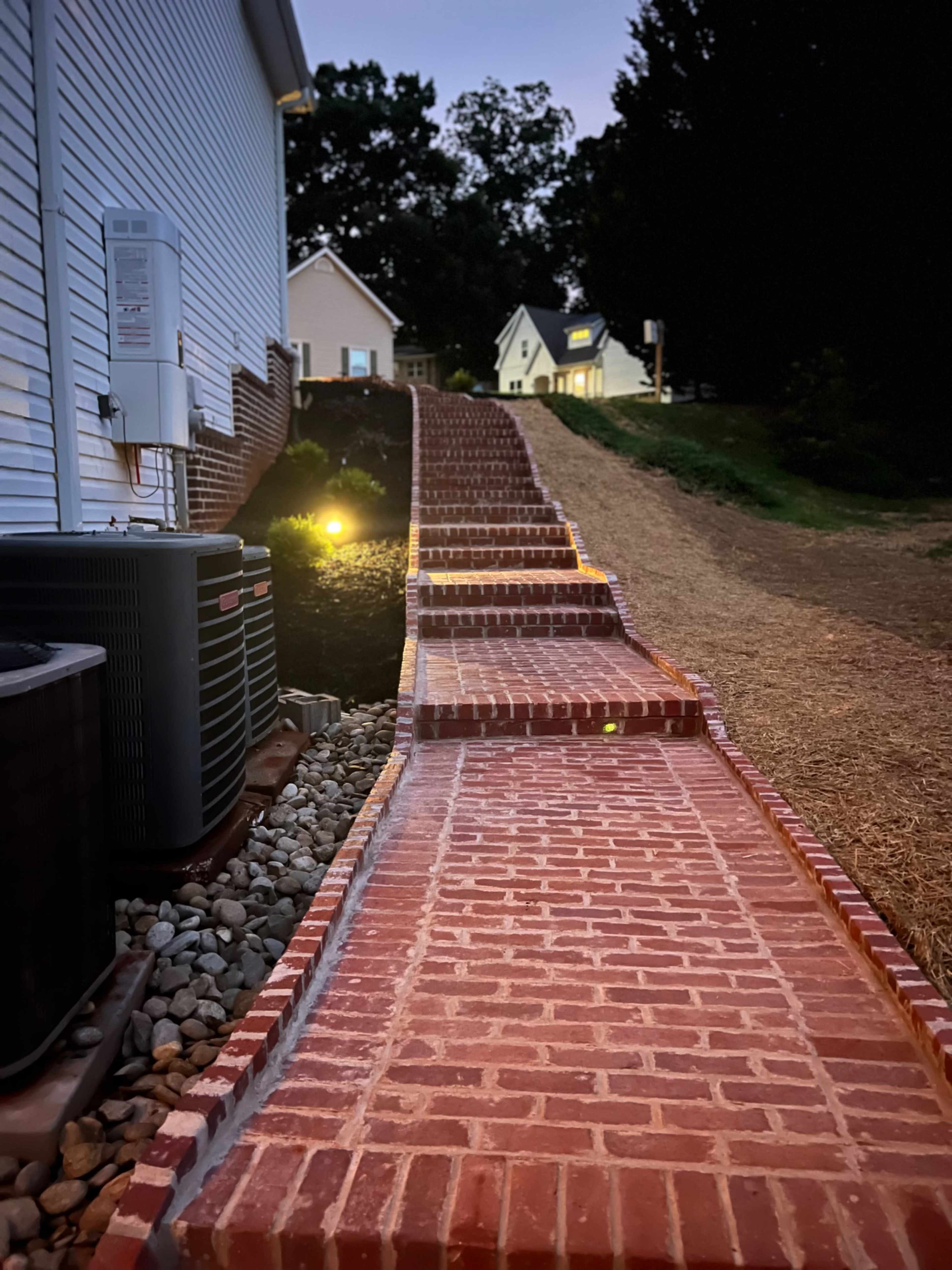 A brick pathway curves upward along a hillside, flanked by grass and a few houses in the distance.