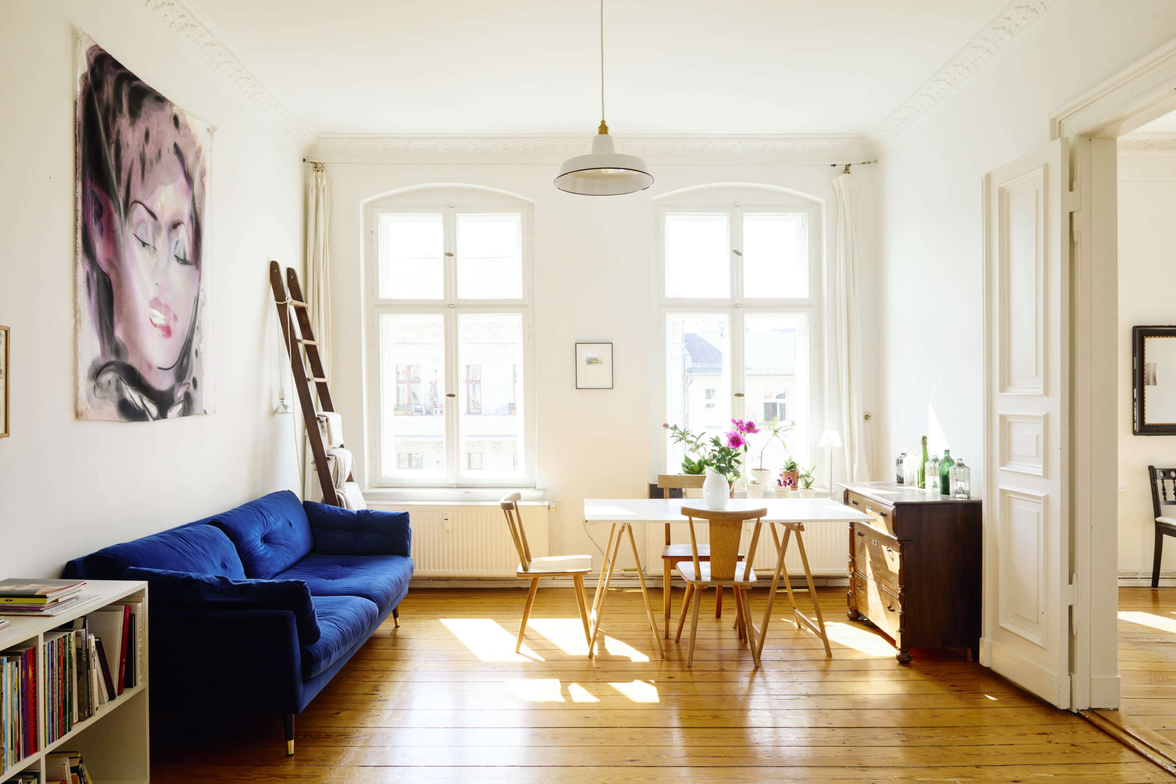 A well-lit living room features a blue sofa, a wooden dining table with chairs, and a painting on the wall, complemented by large windows and wooden flooring.