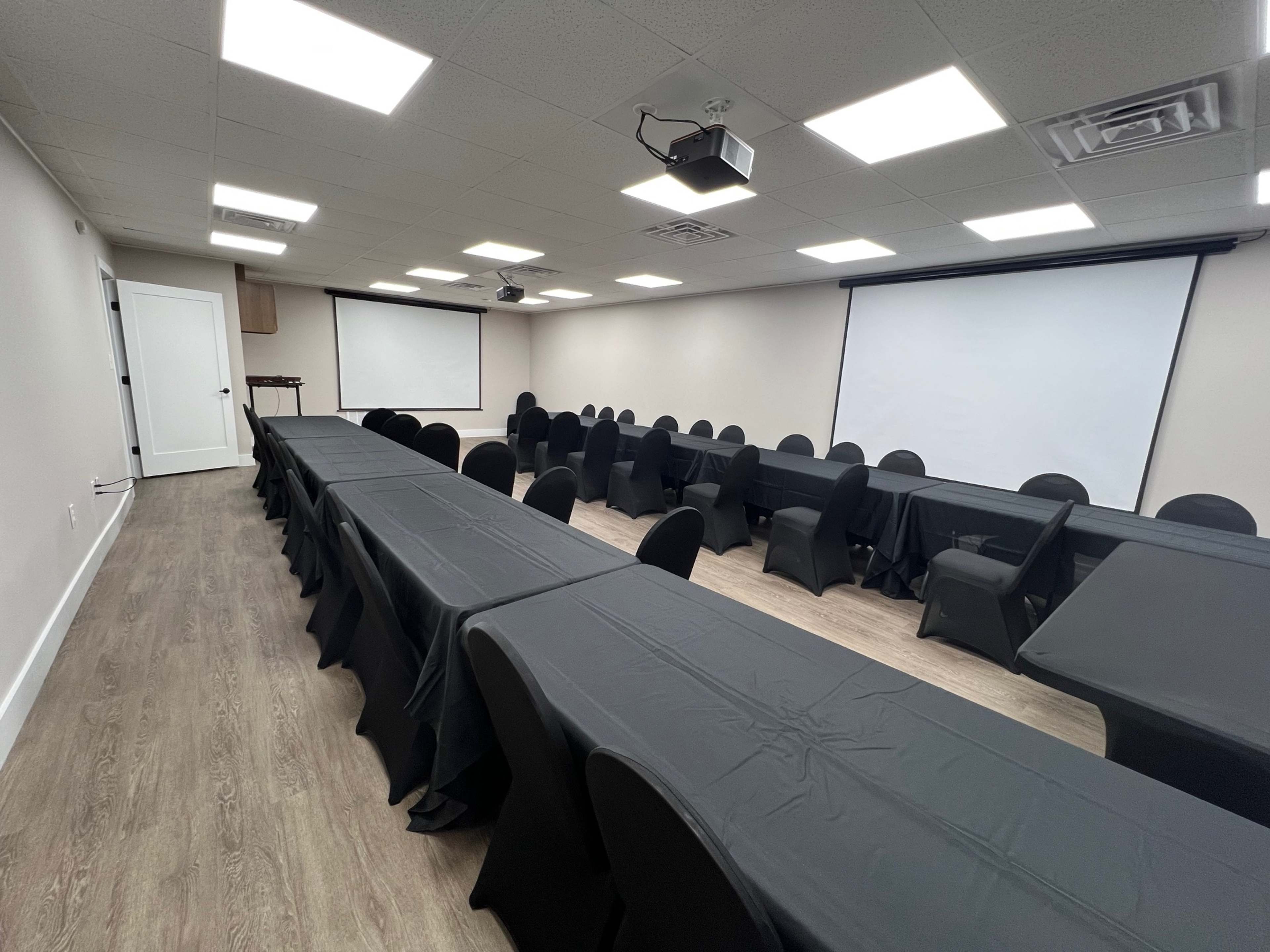 The image shows a conference room set up with multiple long tables covered in black tablecloths and black chairs arranged around them, facing two projection screens.