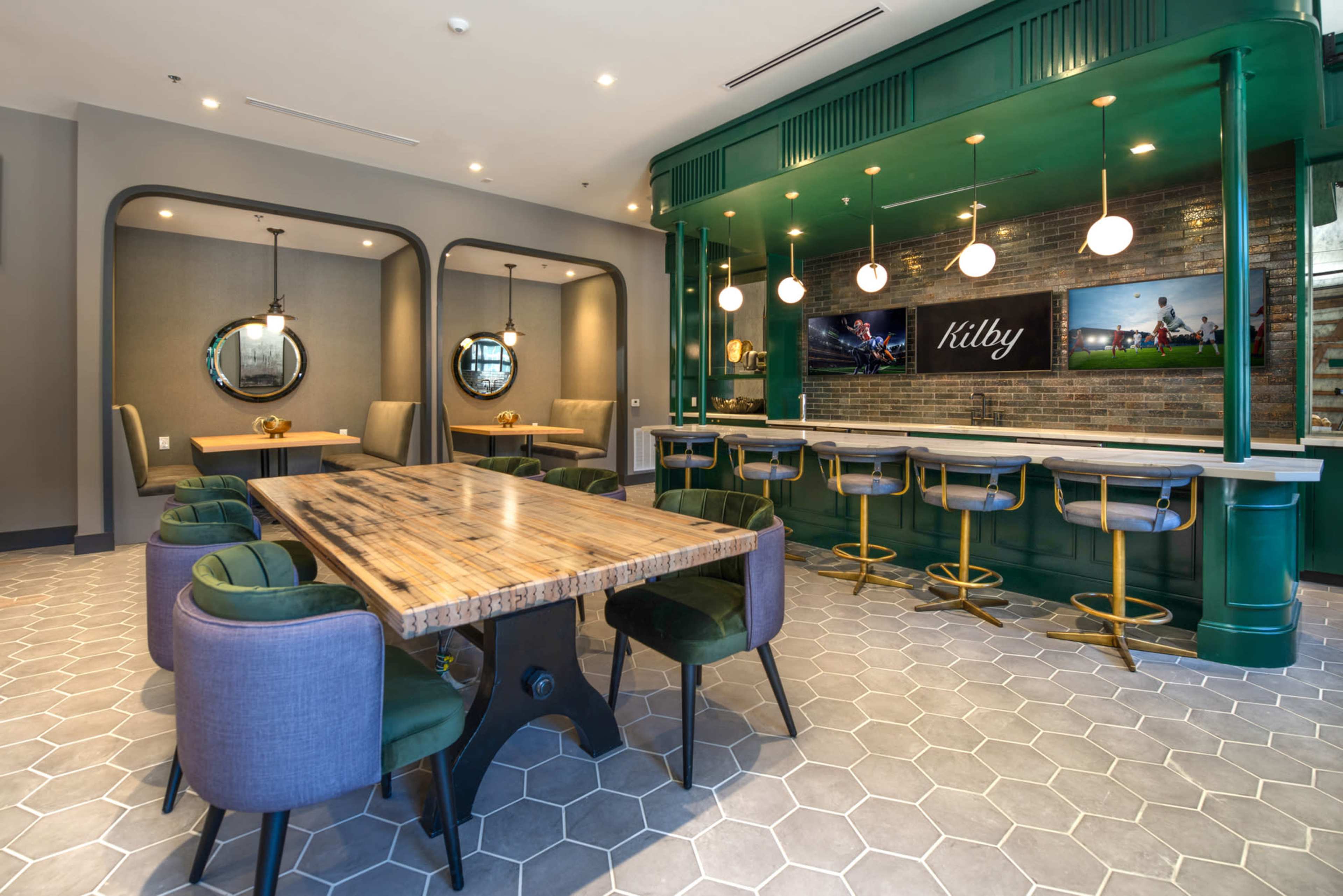 A modern dining area featuring a large wooden table surrounded by upholstered chairs and a bar with green accents and pendant lighting.