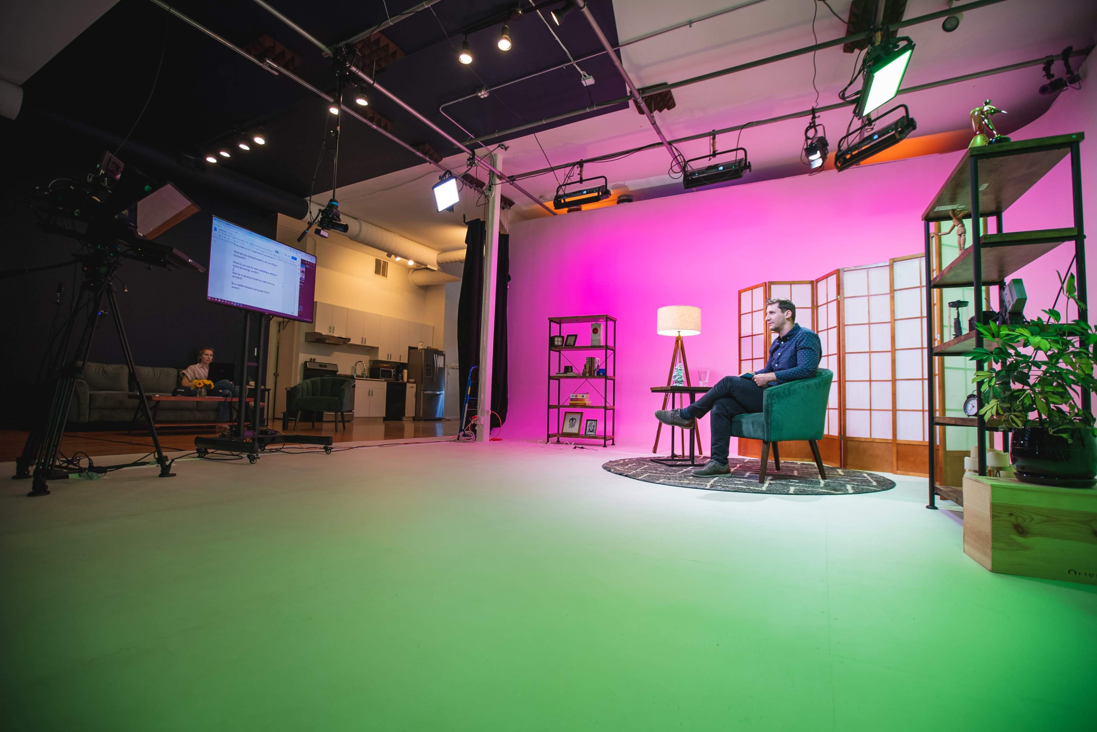 A man sits in a chair on a brightly lit set with a pink backdrop and a plant beside him, while cameras and lighting equipment are positioned around the space.