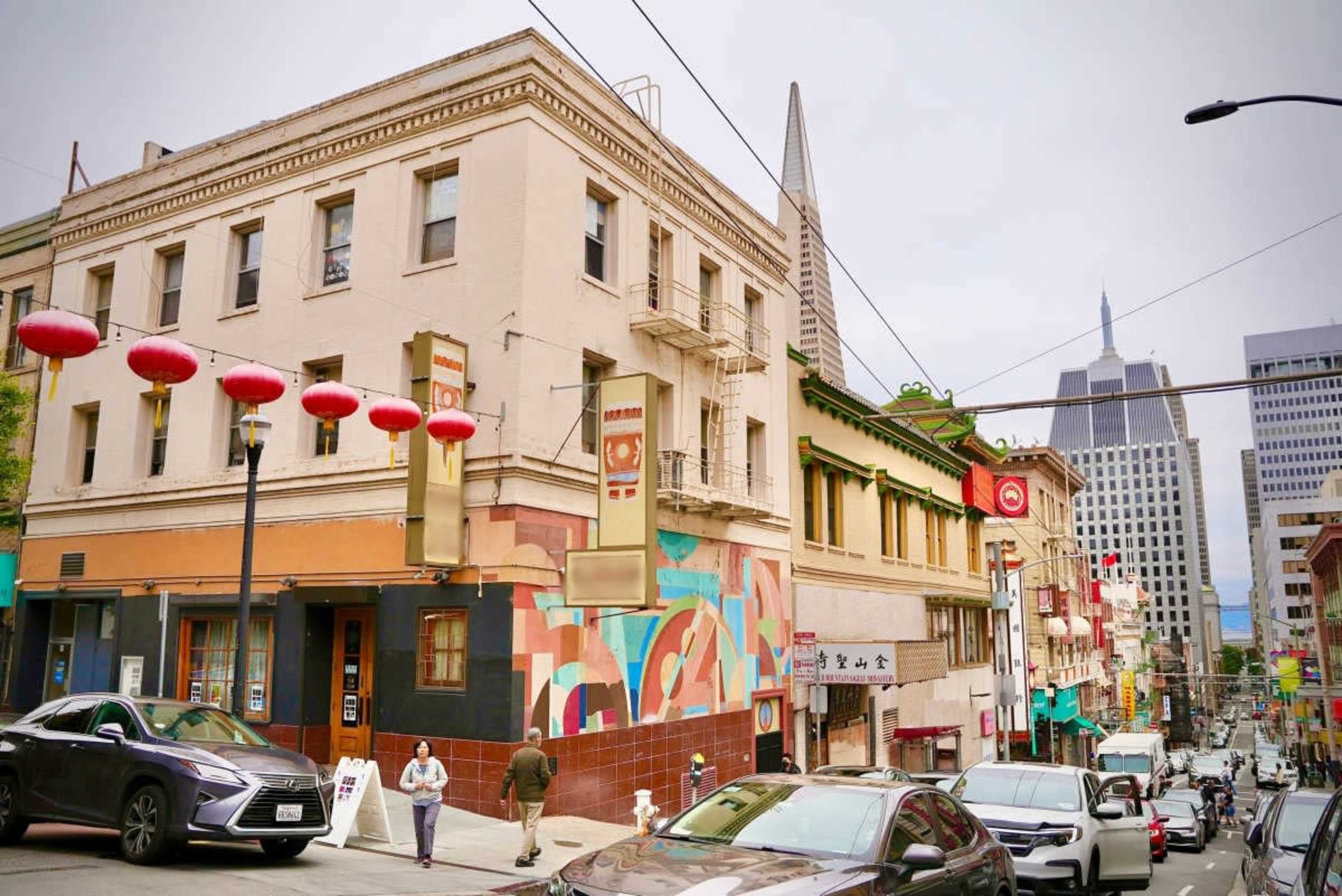 A street scene in a city area featuring a mix of buildings with colorful murals, red lanterns, and vehicles parked along the road.