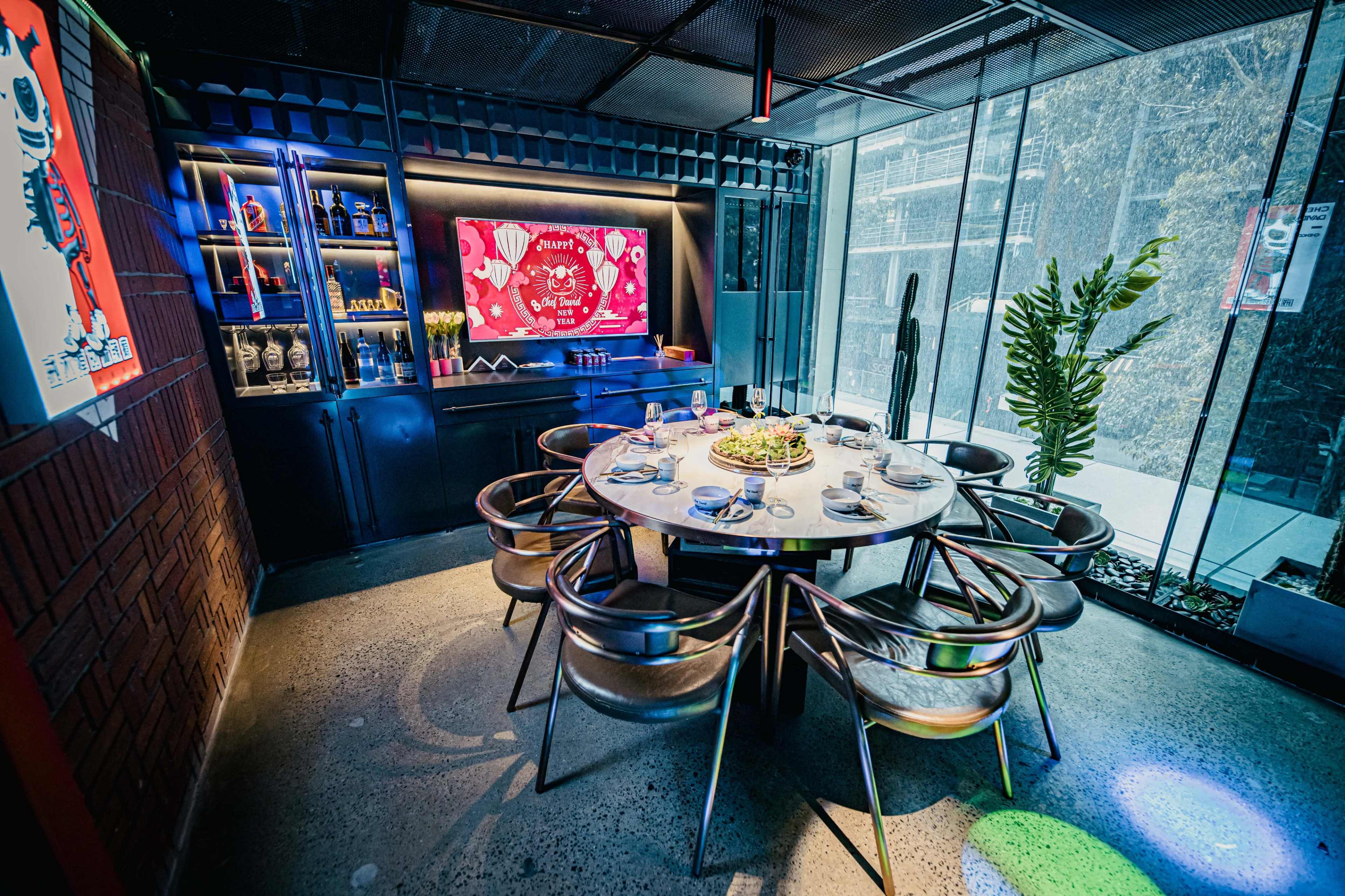 A round dining table is set with plates and cutlery in a modern restaurant featuring colorful lighting and a large screen on the wall.
