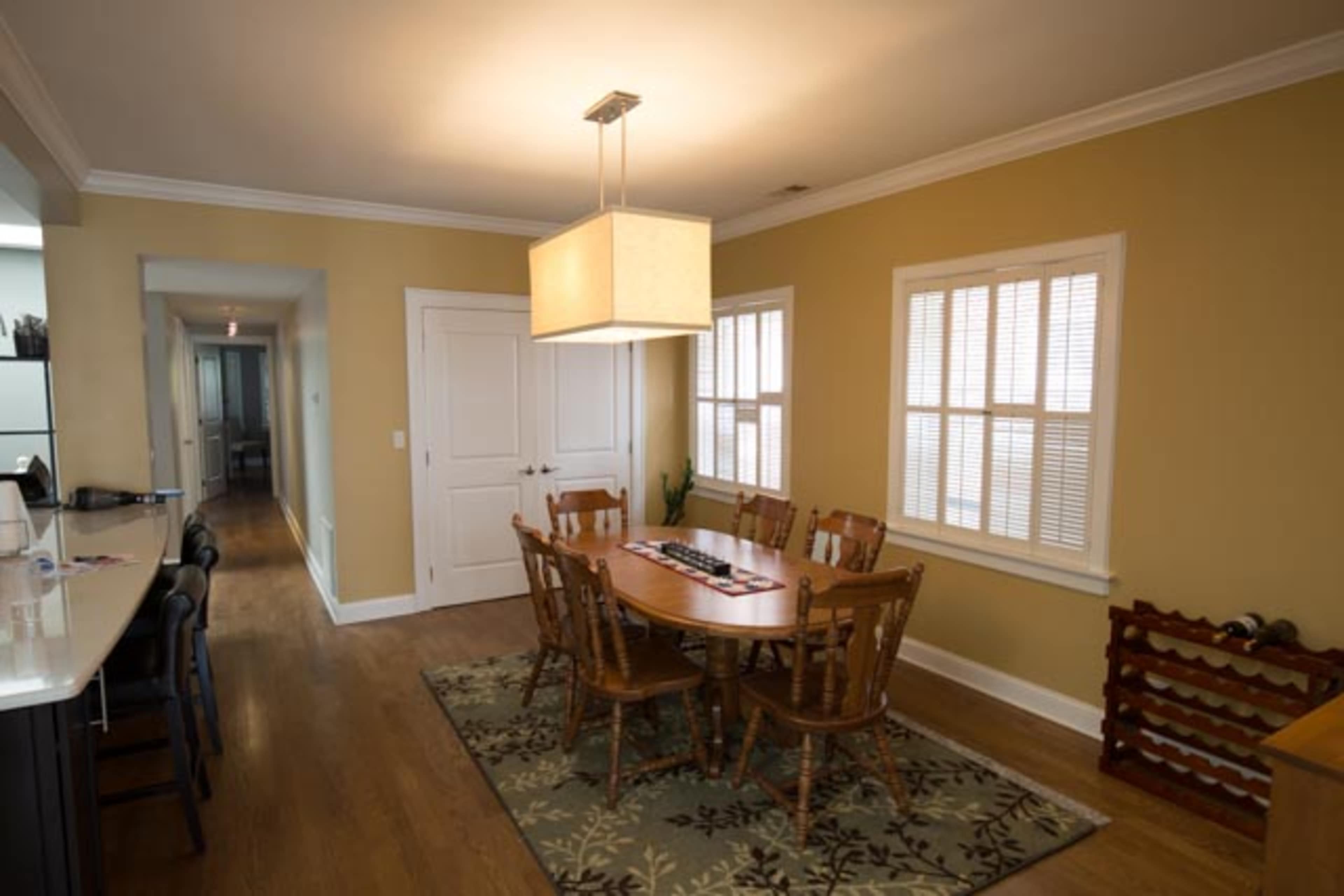 A dining area features a round wooden table surrounded by six chairs, illuminated by a rectangular pendant light, with two windows and a hallway visible in the background.