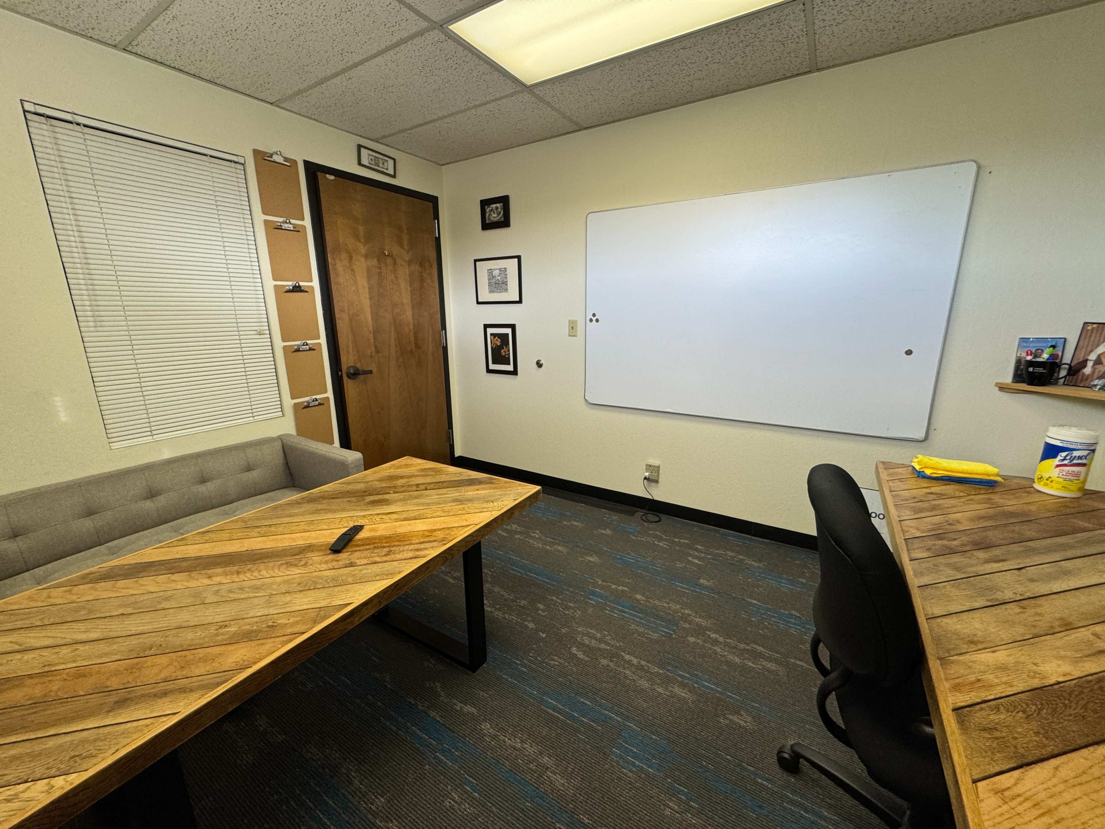 The image shows a small office space with two wooden tables, a whiteboard on one wall, a couch, and a chair.