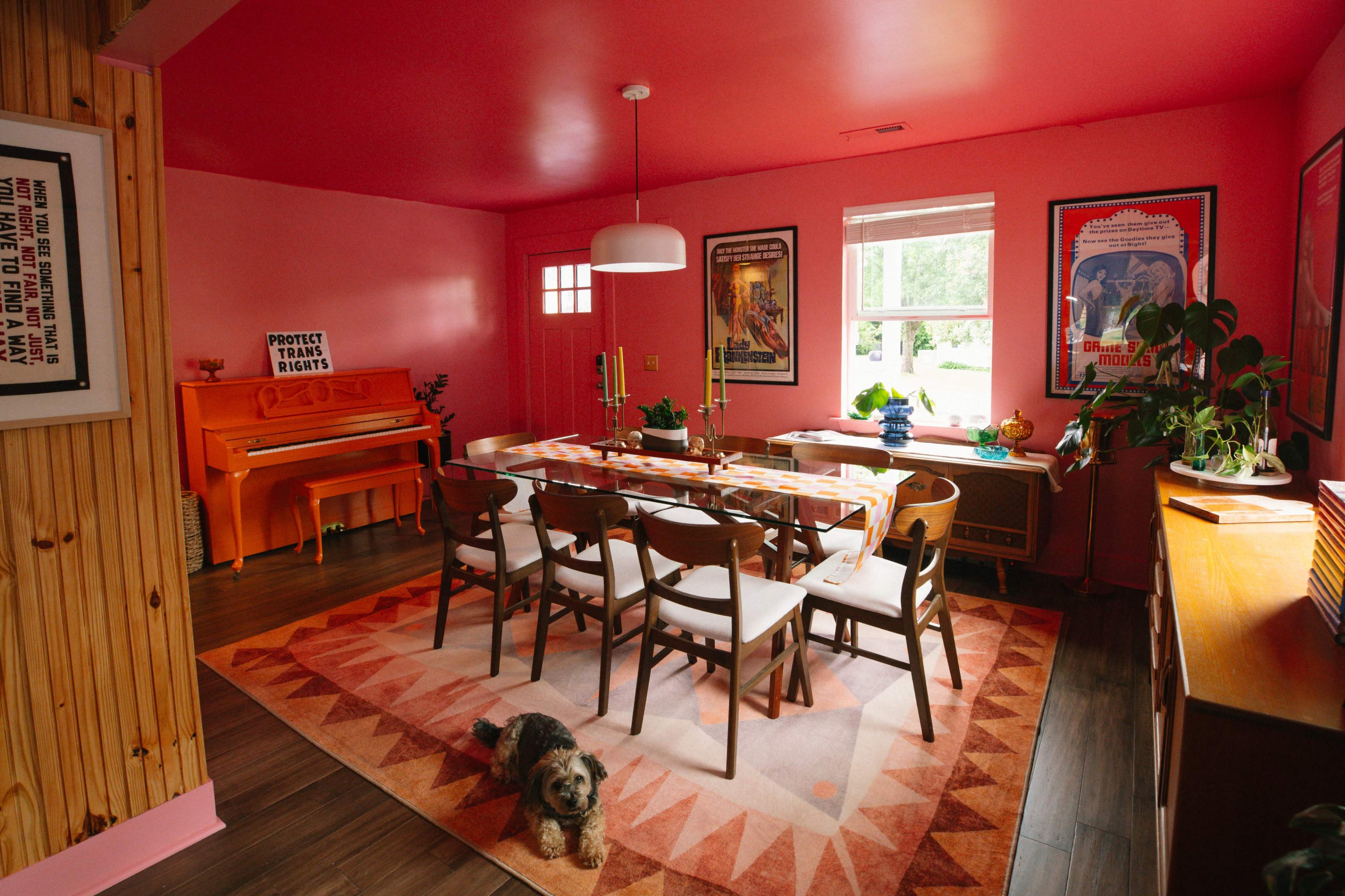 The image shows a vibrant pink dining room featuring a large wooden table surrounded by chairs, an orange piano in one corner, and a dog on a patterned rug.