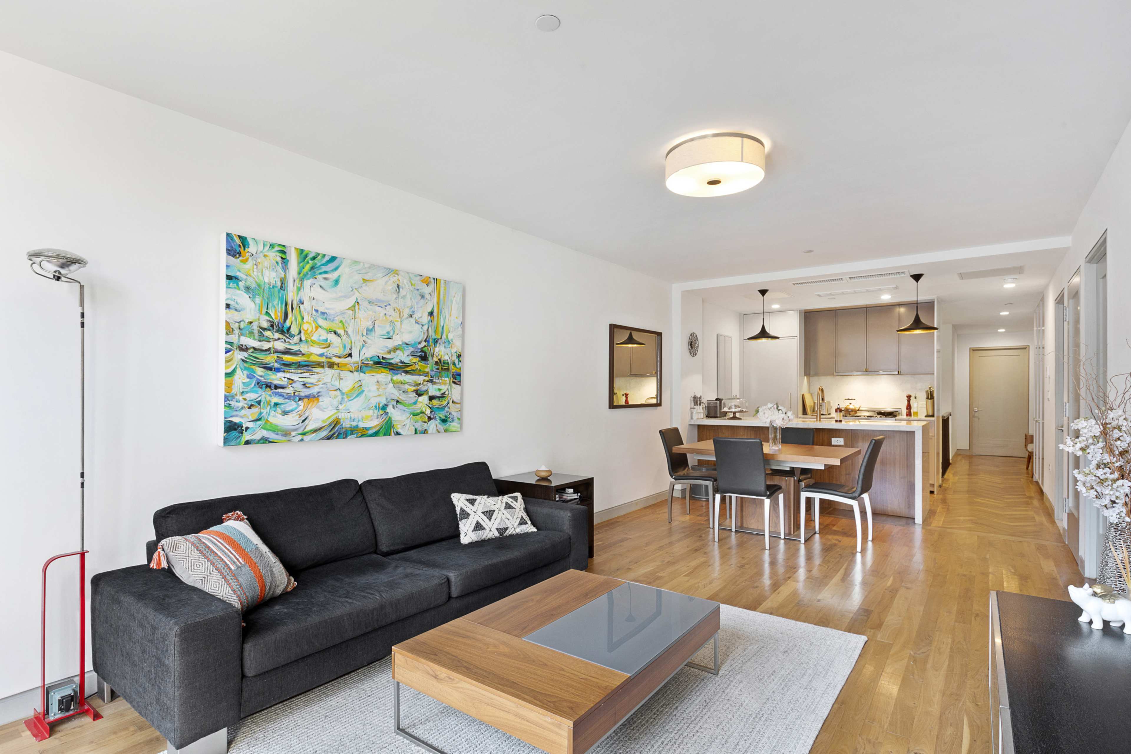 A modern living room with a dark sofa, a coffee table, and a colorful painting on the wall, leading into a dining area with a wooden table and kitchen in the background.