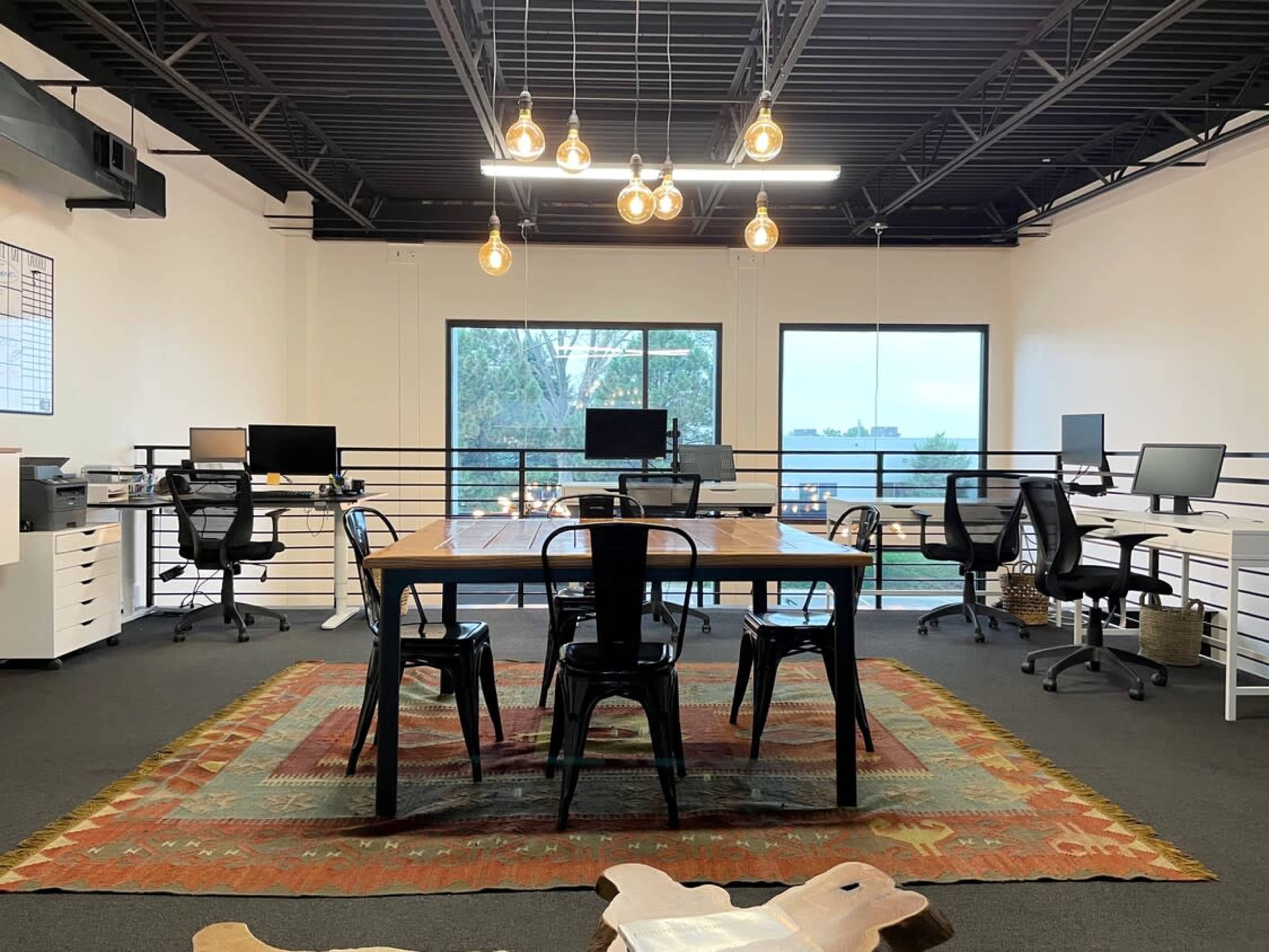 A modern office space features a large wooden table surrounded by chairs, with multiple computer workstations and large windows overlooking greenery.